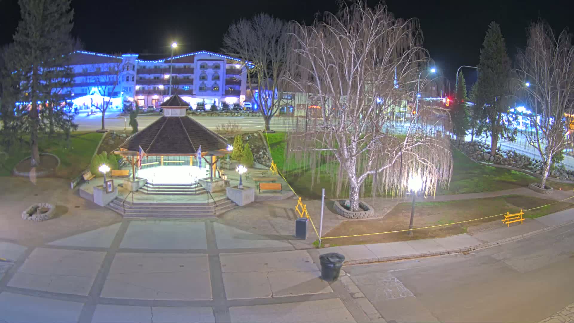 A nighttime view of a town square featuring a gazebo, park, and a multi-story building, illuminated by streetlights.