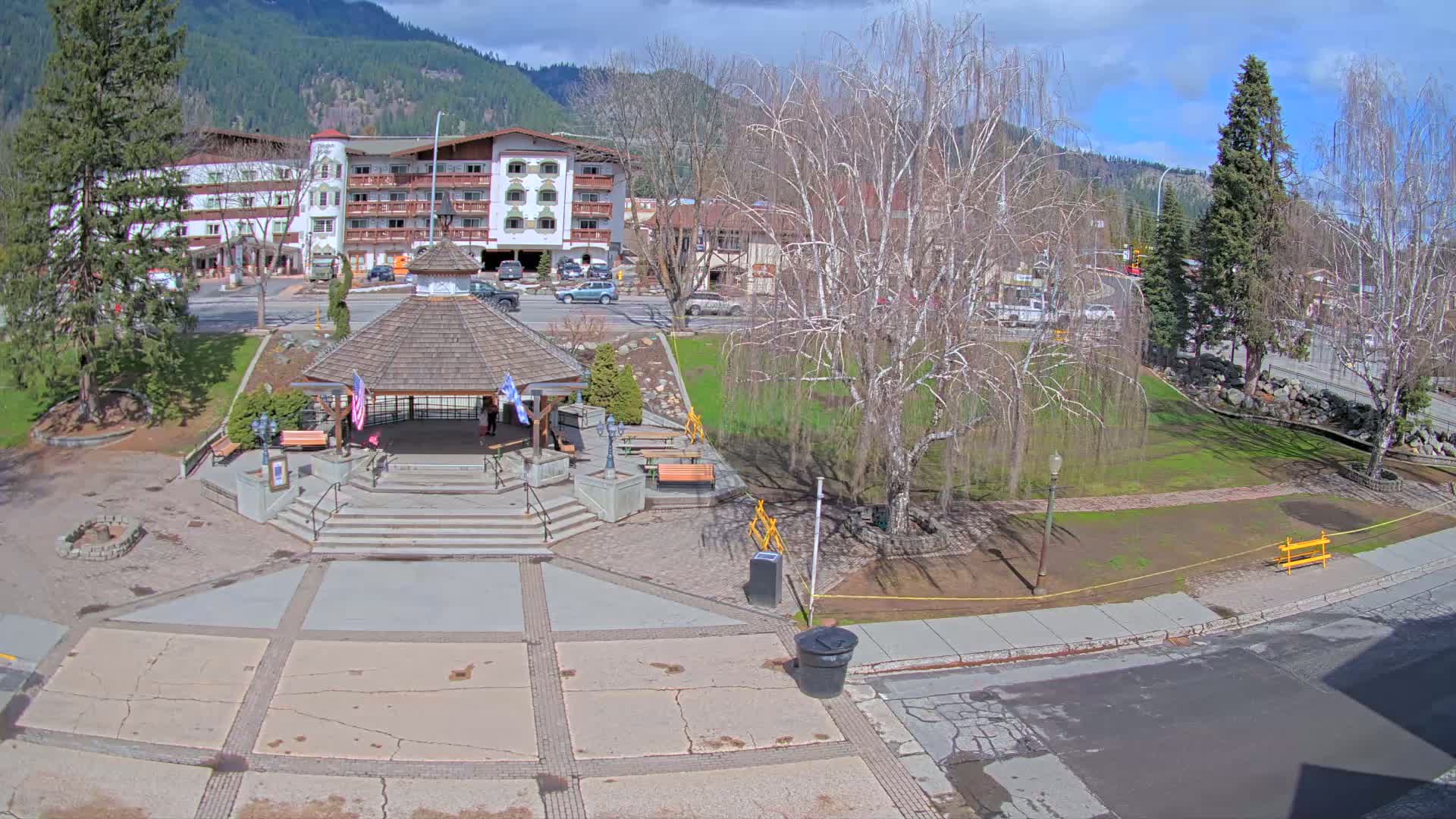 A paved plaza with a gazebo and stairs, a grassy park with bare trees, and a multi-story building in the background, all under a mostly sunny sky.