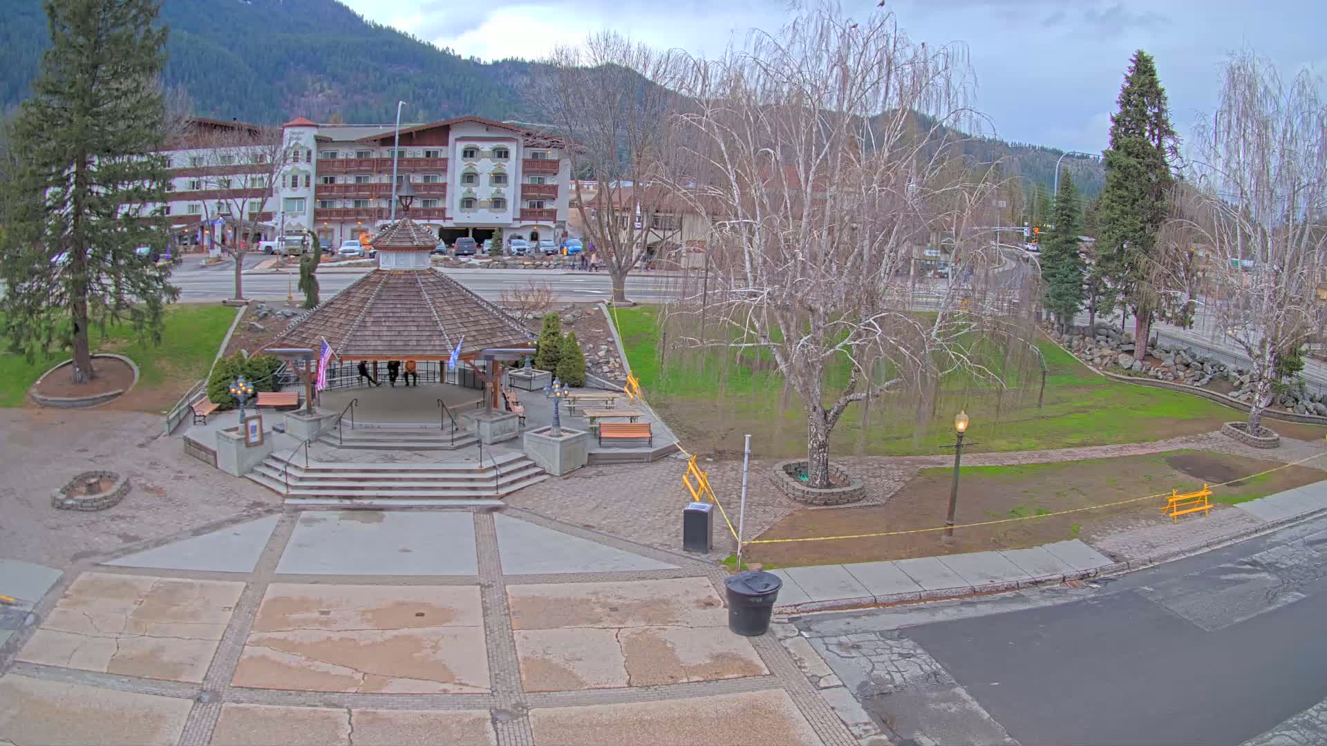 A park with a gazebo, picnic tables, and a large tree is situated in front of a multi-story building, under a mostly cloudy sky.