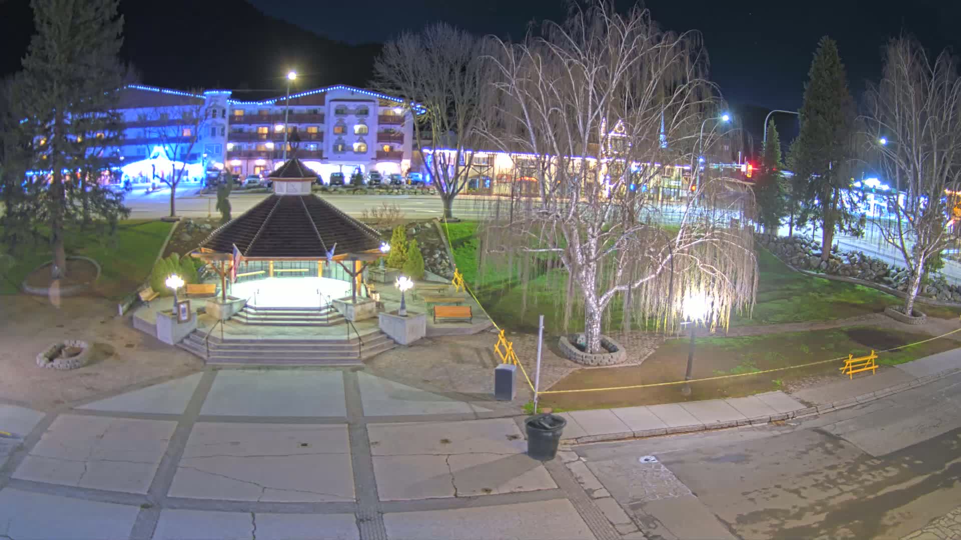 A nighttime view of a town square with a gazebo, trees, and a multi-story building illuminated with lights.