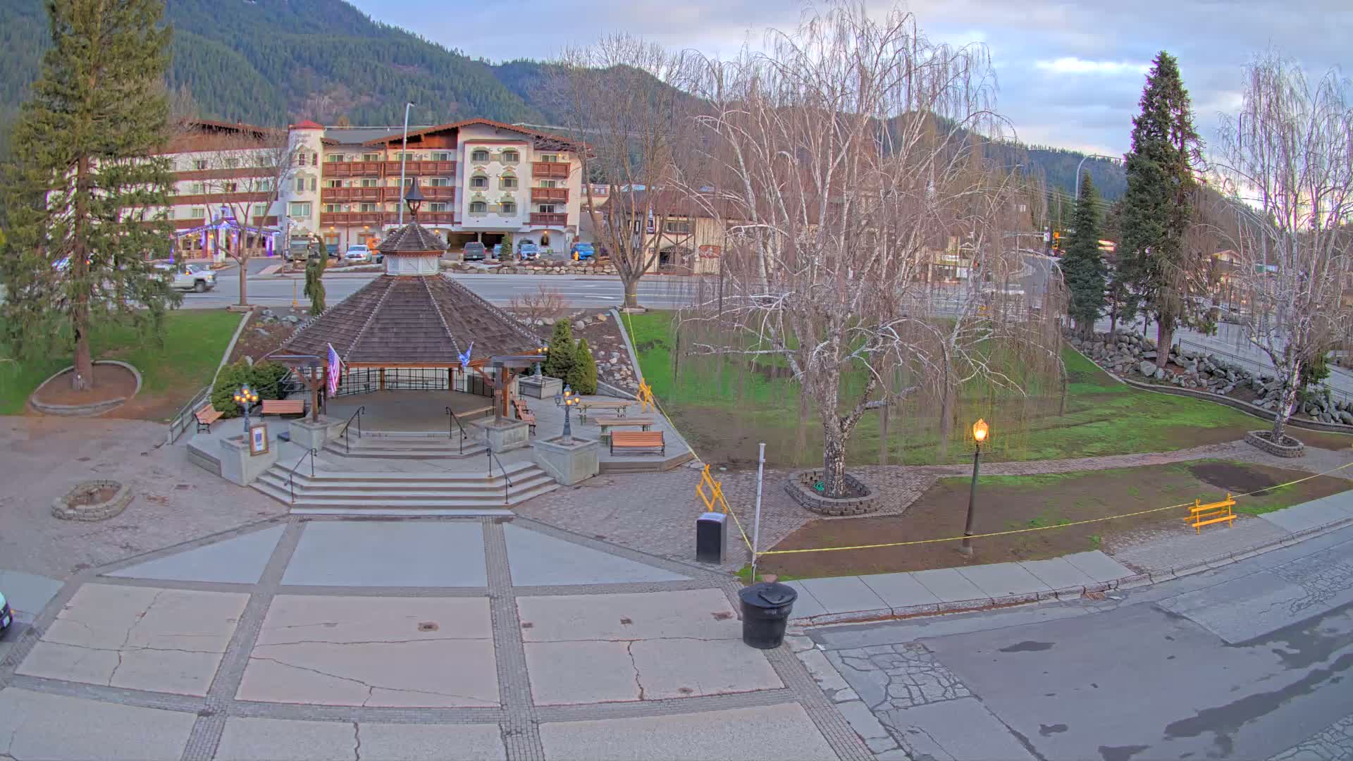 A gazebo with steps sits in a town square, next to a grassy area with bare trees, under a cloudy sky, with a hotel in the background and mountains beyond.