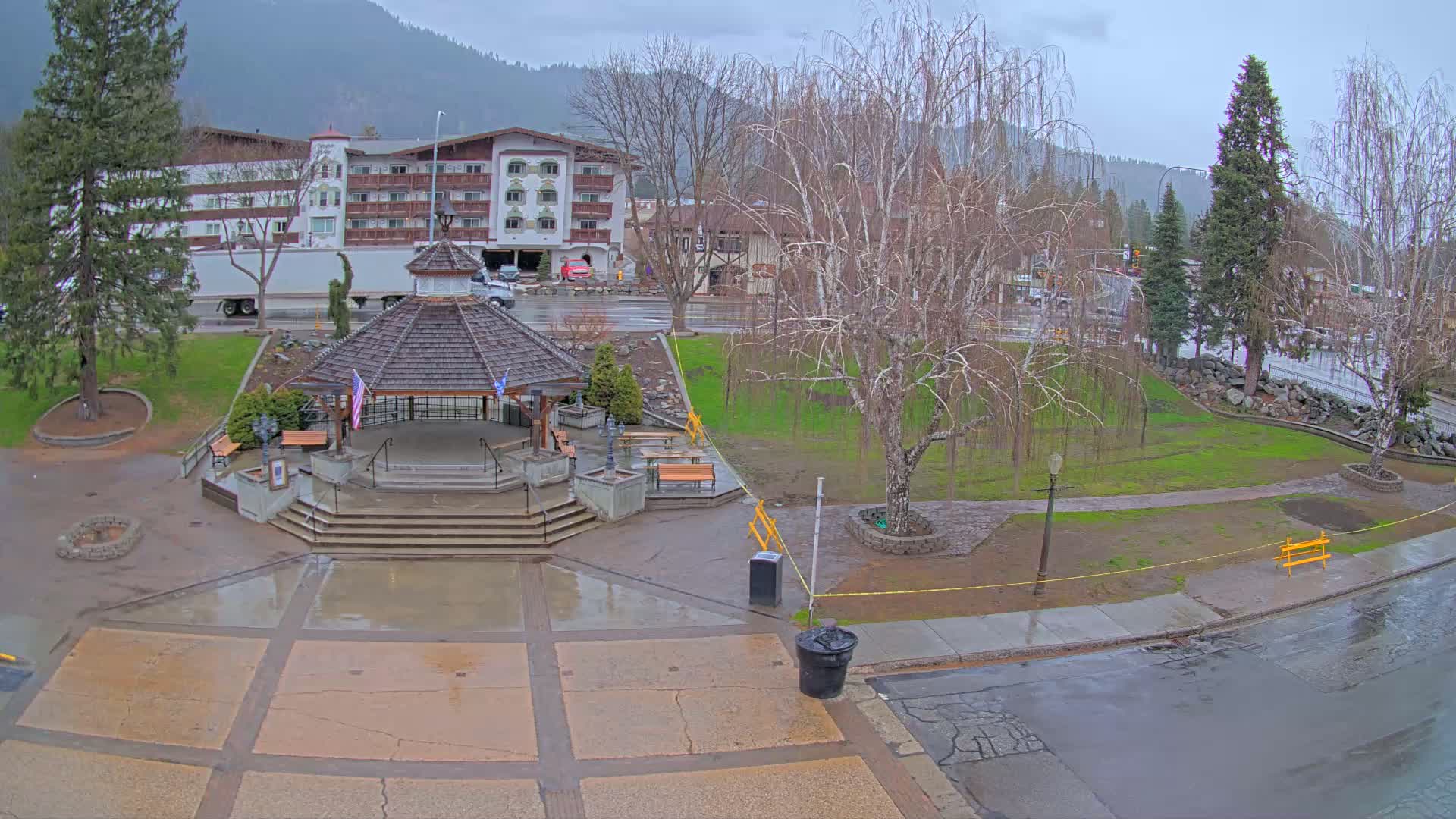 A wet town square with a gazebo and benches sits under a cloudy sky, with a multi-story building and trees in the background.