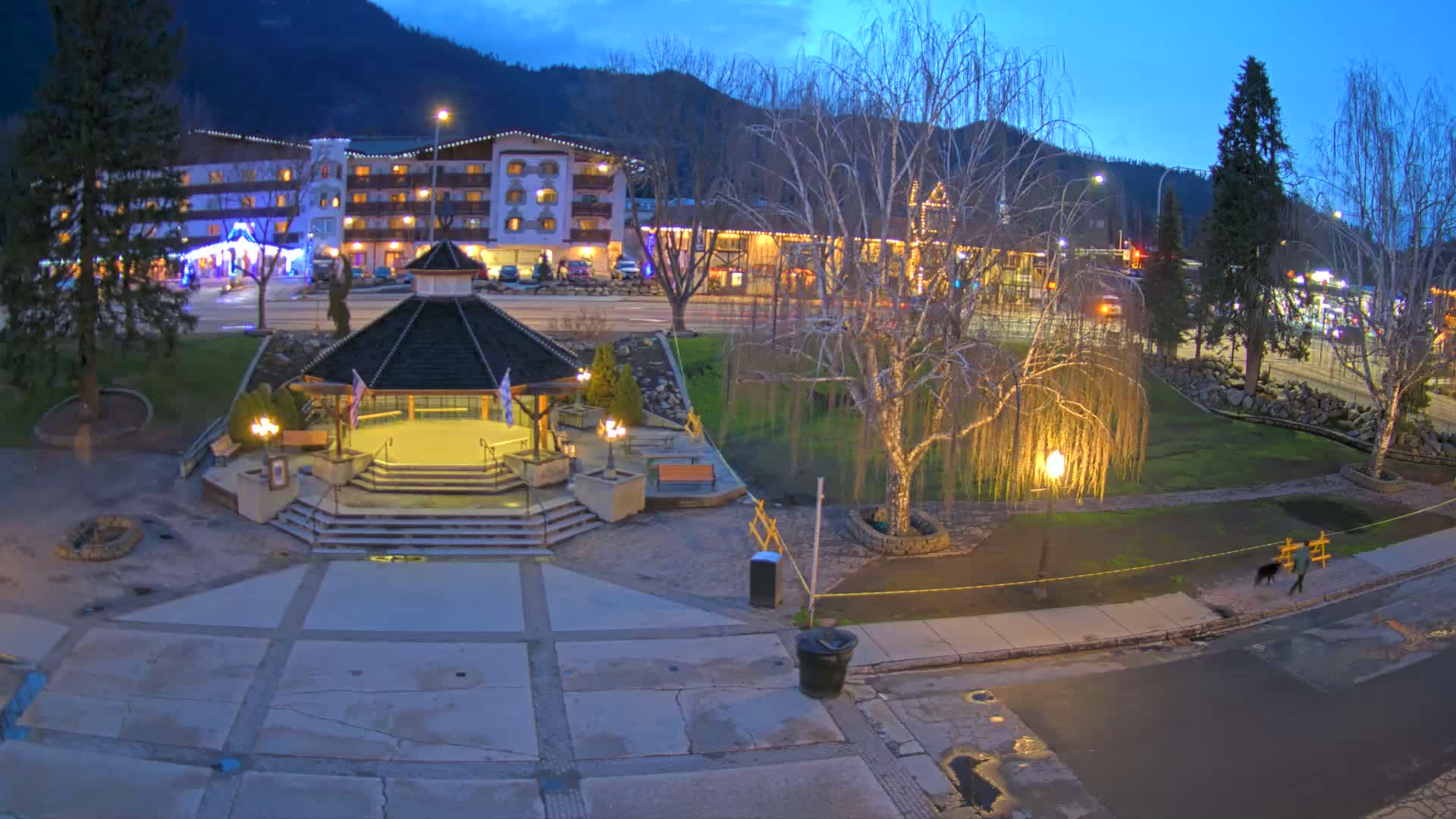 A gazebo sits in a town square at dusk, with a hotel and other buildings visible in the background under a twilight sky.