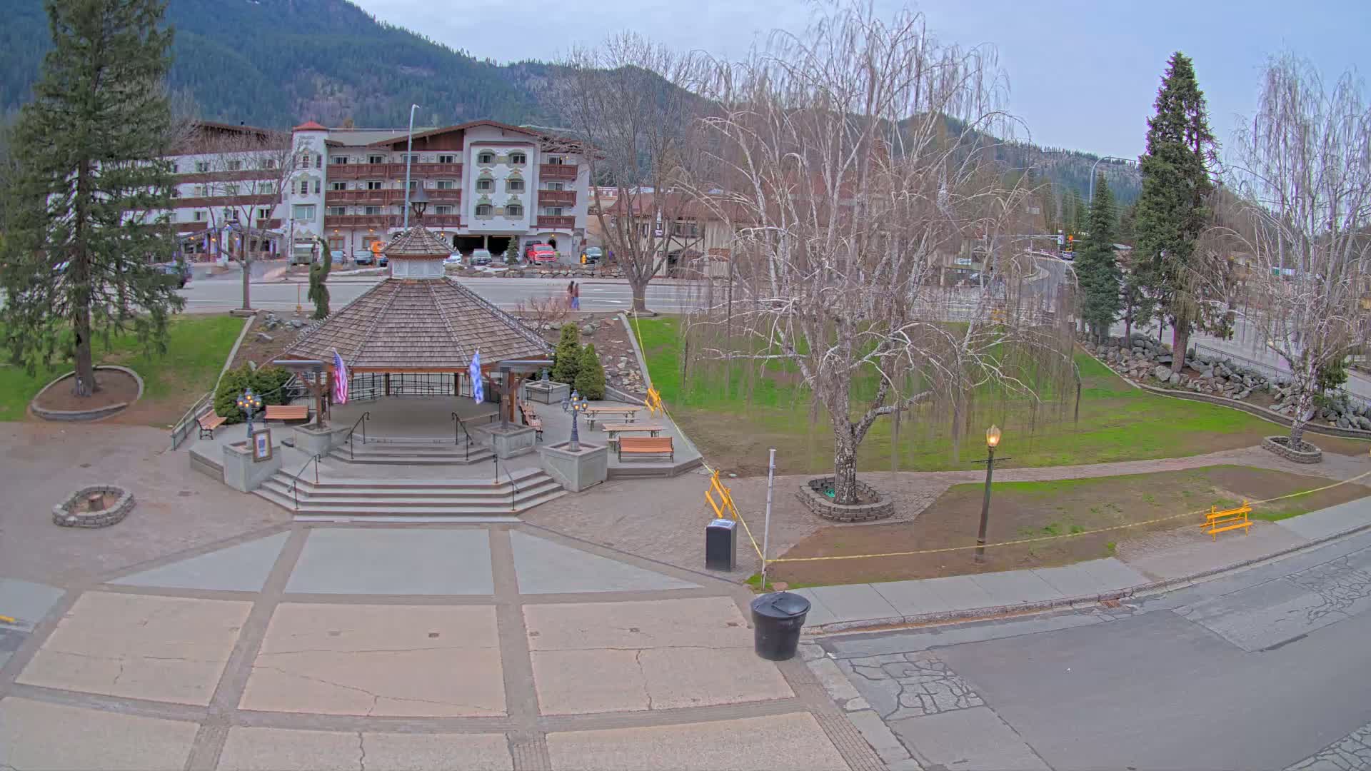 A paved town square with a gazebo, benches, and trees, situated in front of a multi-story building, under a mostly cloudy sky.