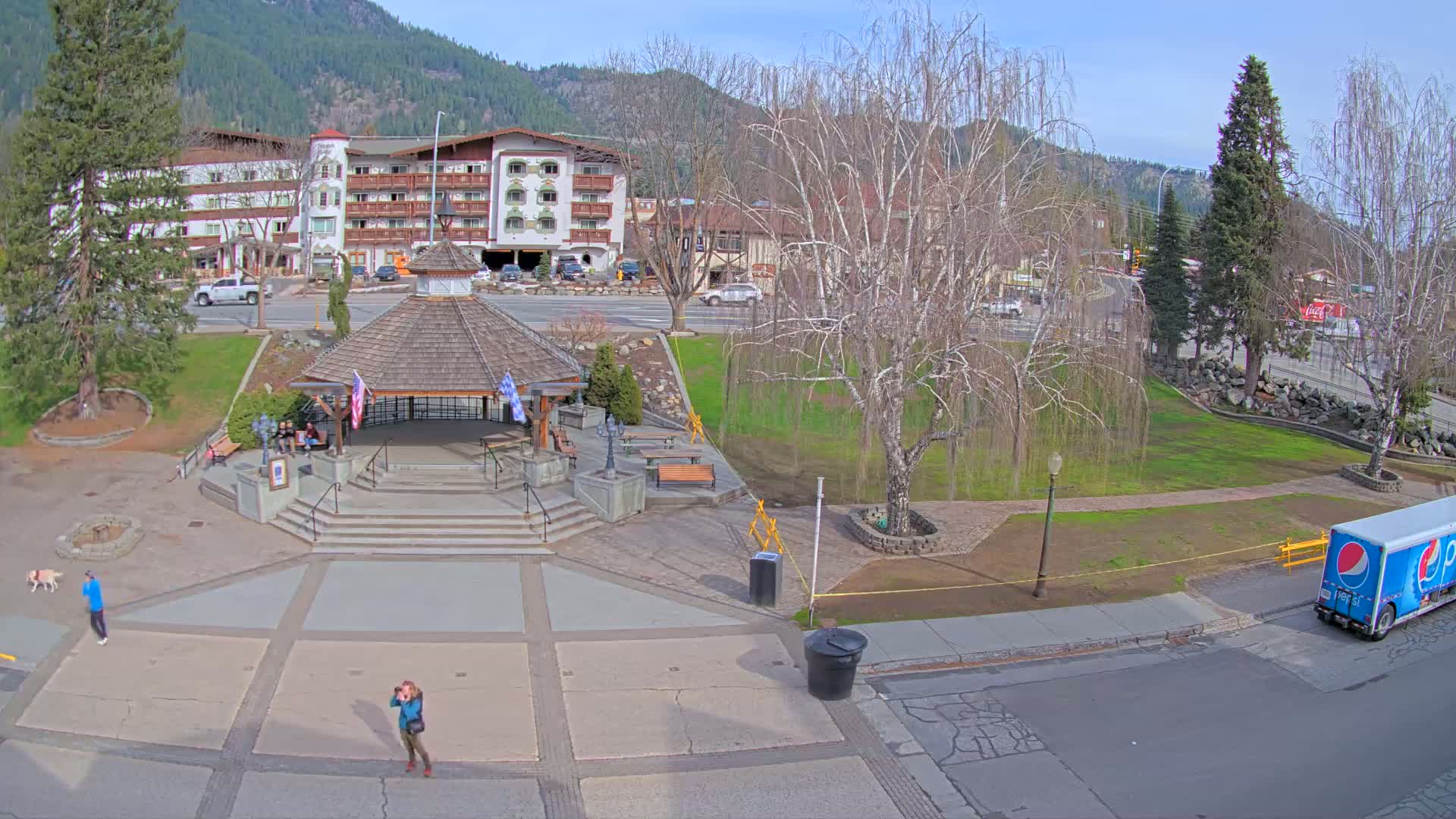 A paved town square with a gazebo, a few people, and a dog, next to a street with a Pepsi truck, under a sunny sky, in front of a multi-story building and mountains in the background.