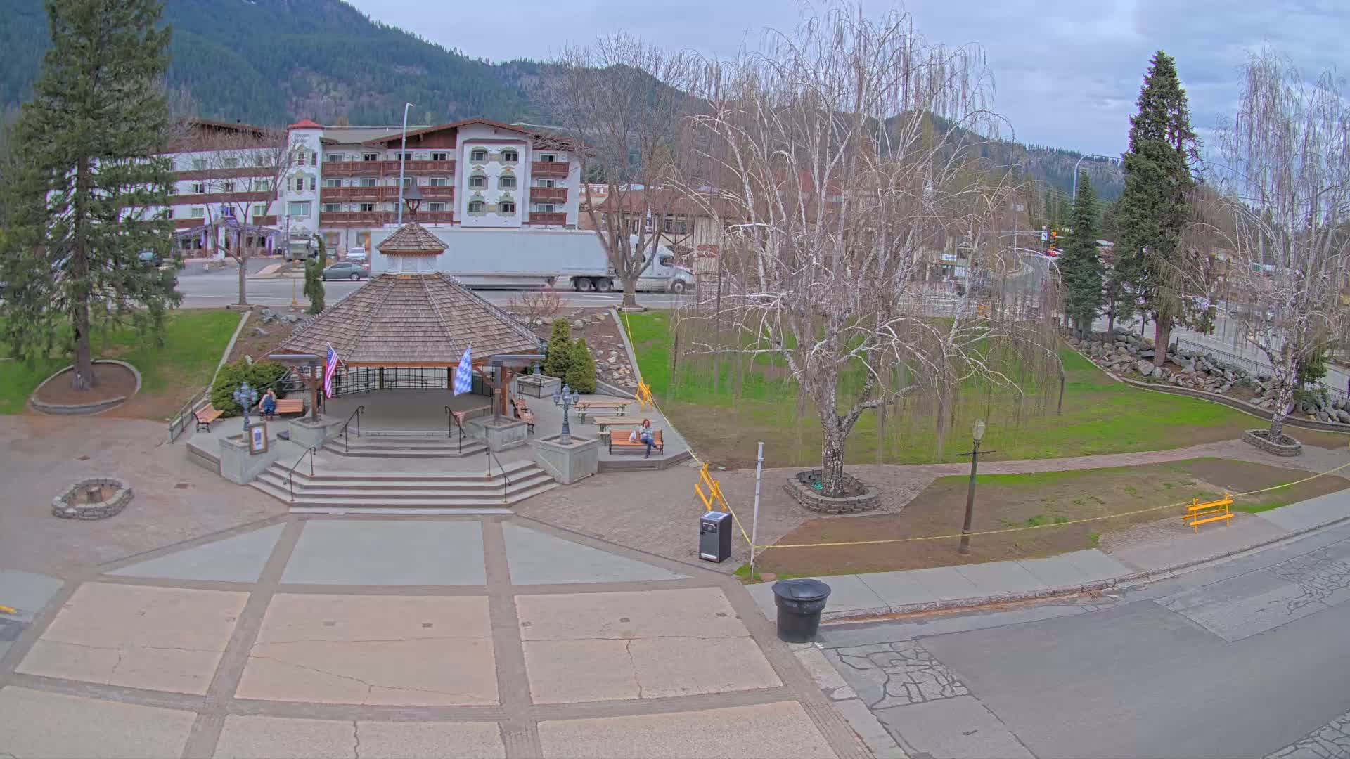 A paved town square with a gazebo, park benches, and a large tree, situated in front of a multi-story building and under a cloudy sky.