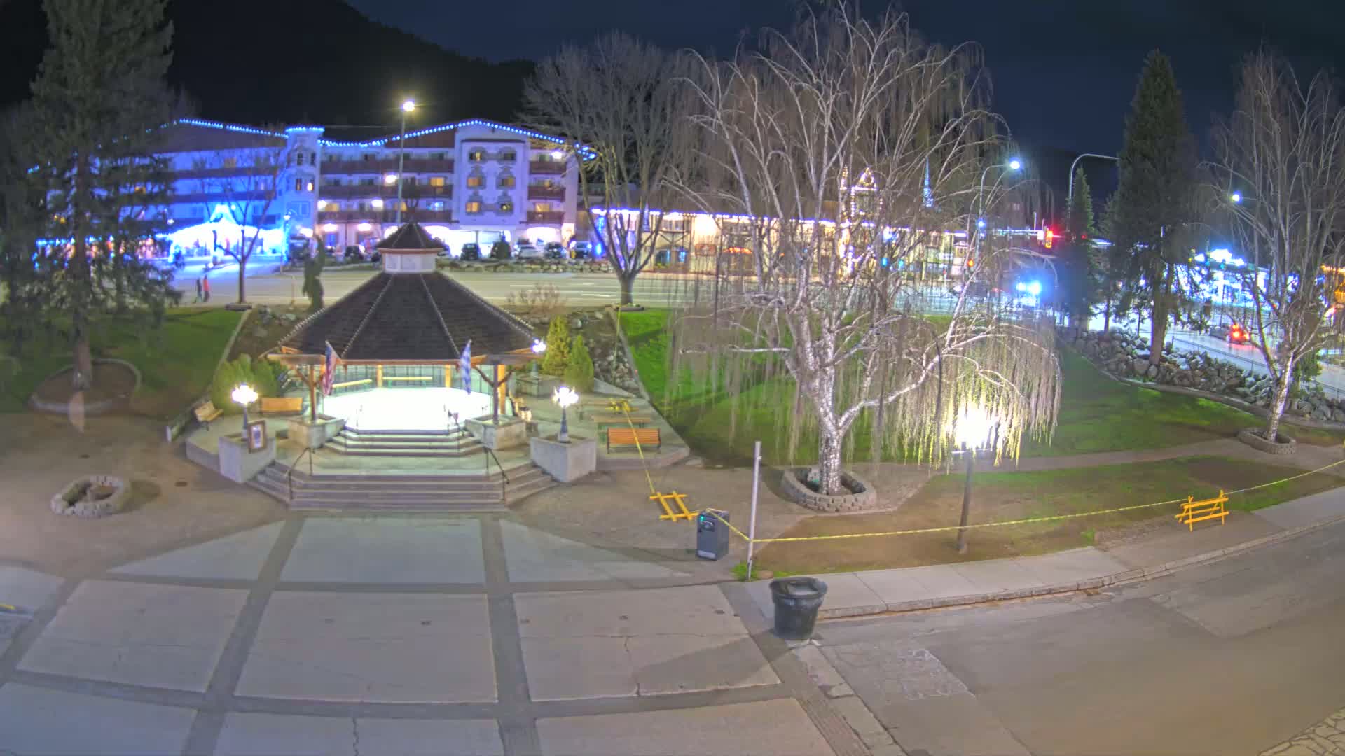 A nighttime view of a town square with a gazebo, trees, and a multi-story building illuminated with lights.