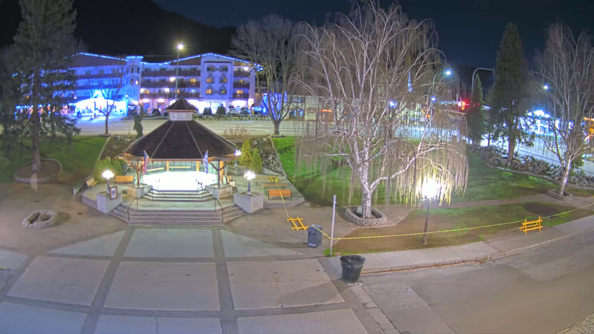 A nighttime view of a town square featuring a gazebo, a park with trees, and a multi-story building in the background.