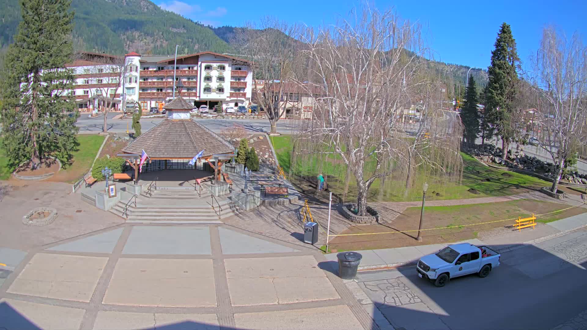A town square with a gazebo, benches, and a large tree, situated in front of a multi-story building, all under a clear, sunny sky with mountains in the background.
