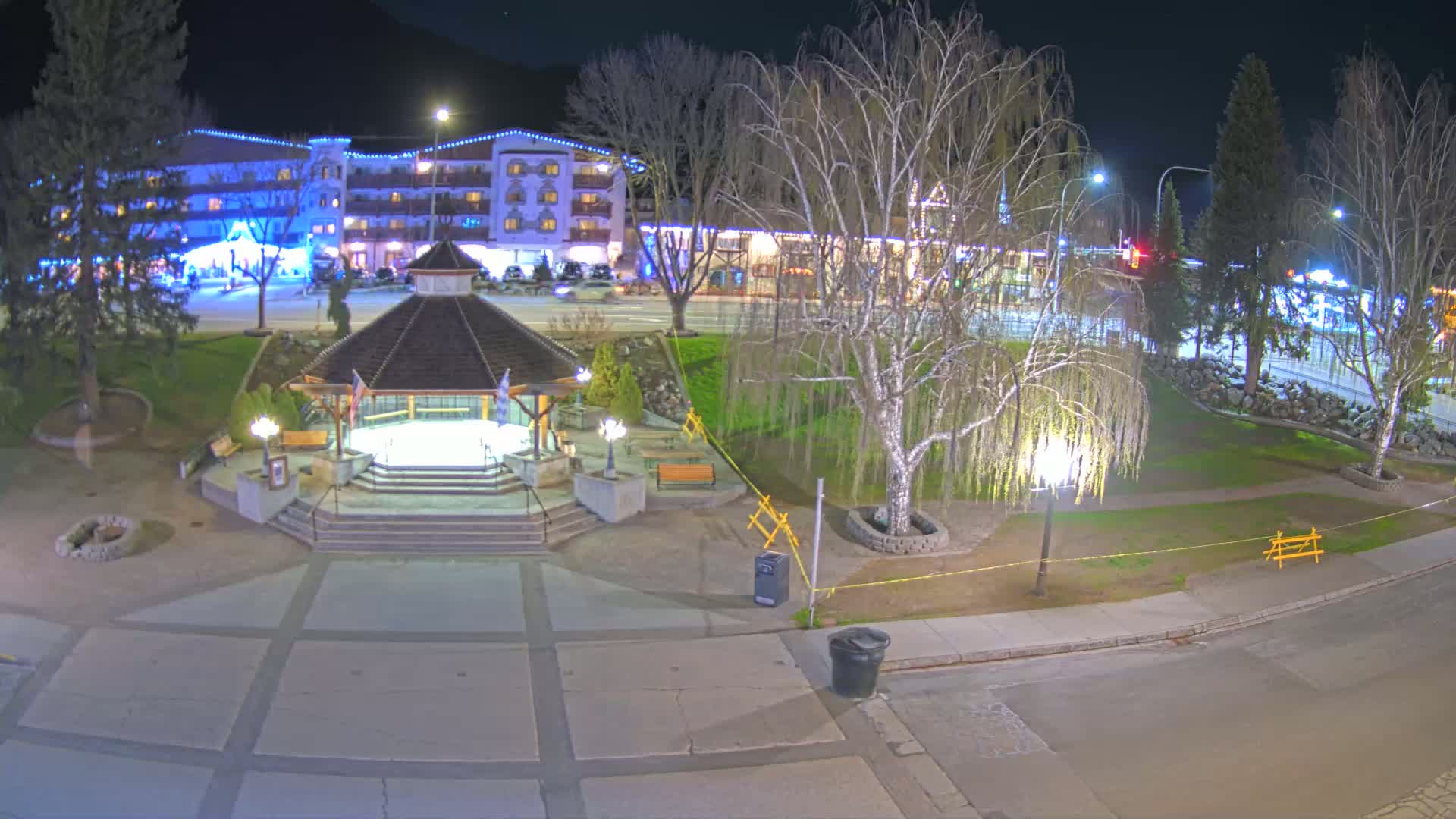 A nighttime view of a town square with a gazebo, park benches, trees, and a building illuminated with string lights.