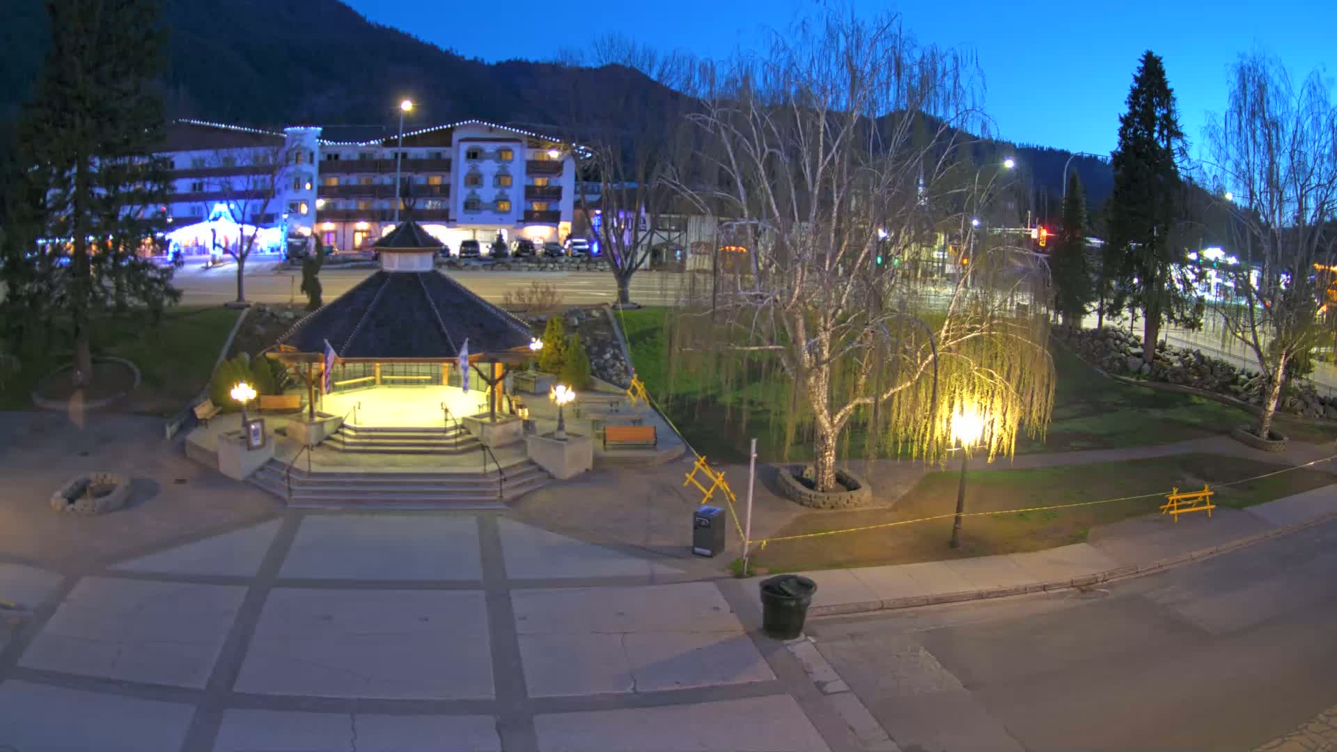 A nighttime view of a town square with a gazebo, trees, and a hotel in the background under a twilight sky.