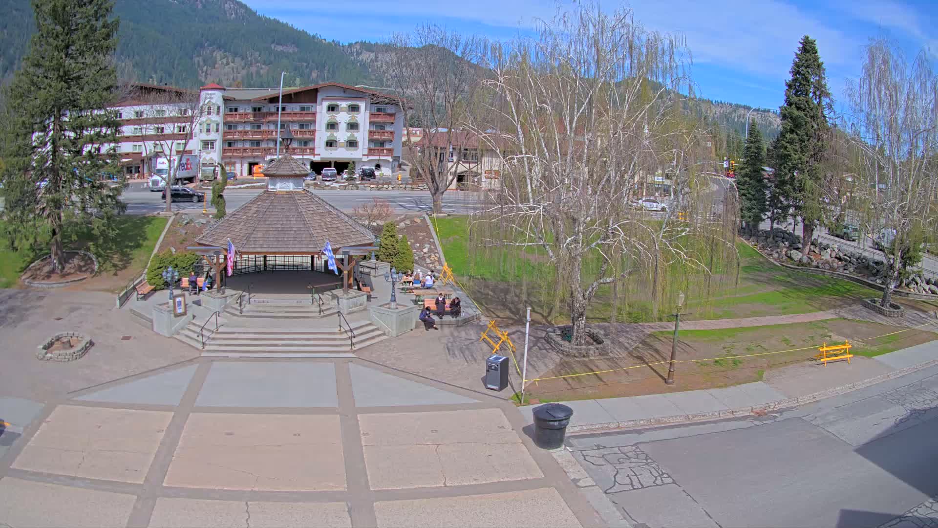A paved plaza with a gazebo and a few people sitting on benches is situated in front of a multi-story building, all under a sunny sky, with mountains in the background.