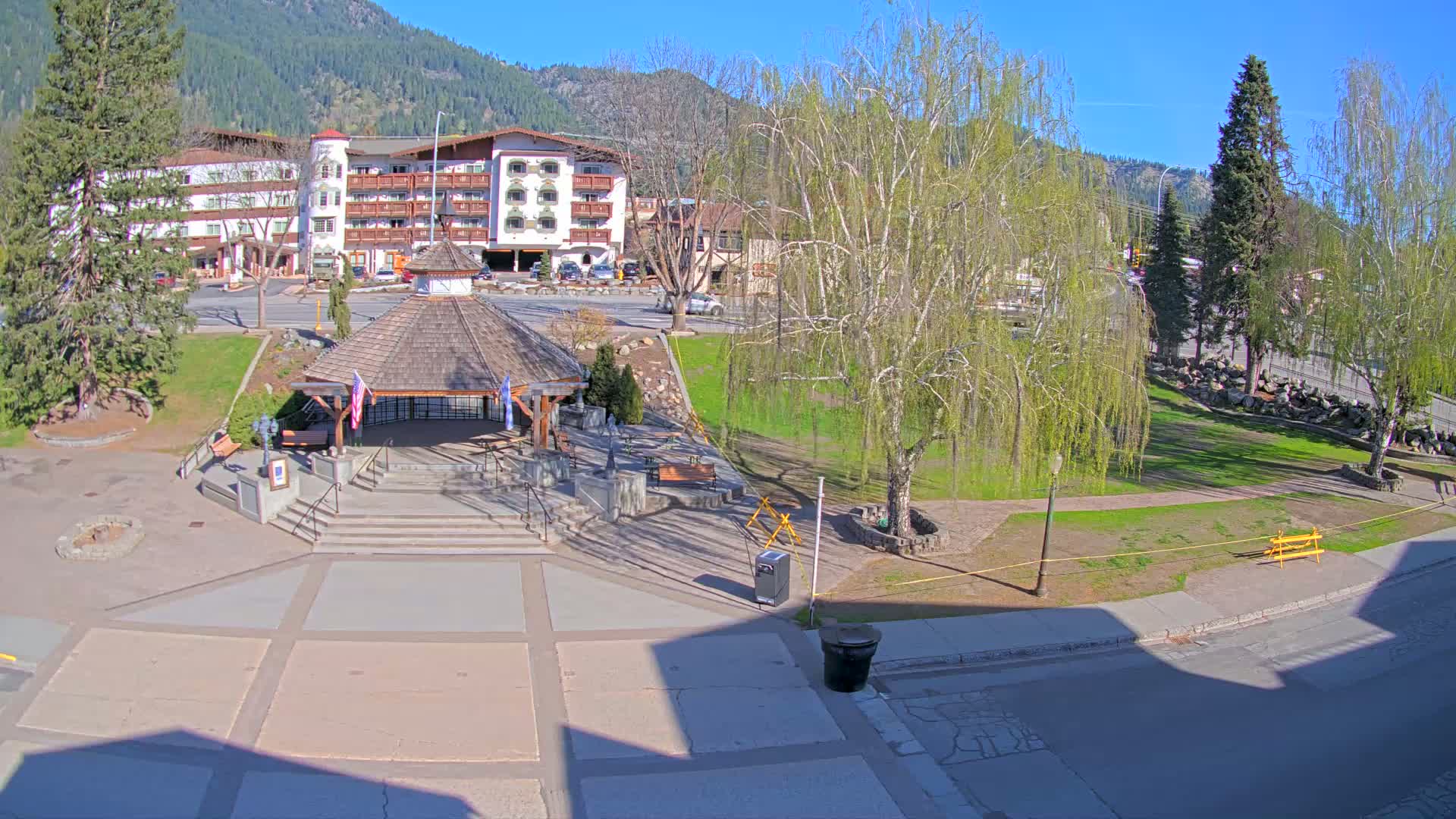 A paved plaza with a gazebo and steps, a grassy park with trees, and a multi-story building in the background on a sunny day.
