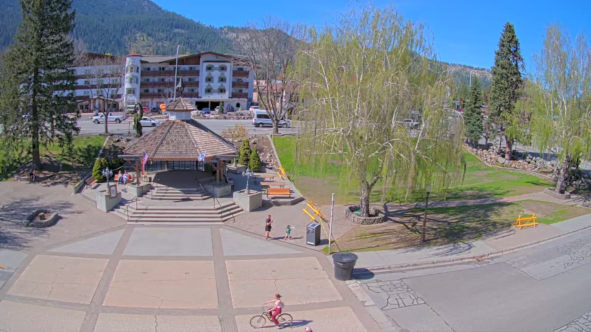 A paved town square with a gazebo, benches, and a few people, including a cyclist, is situated in front of a multi-story building under a sunny sky, with mountains in the background and trees surrounding the square.