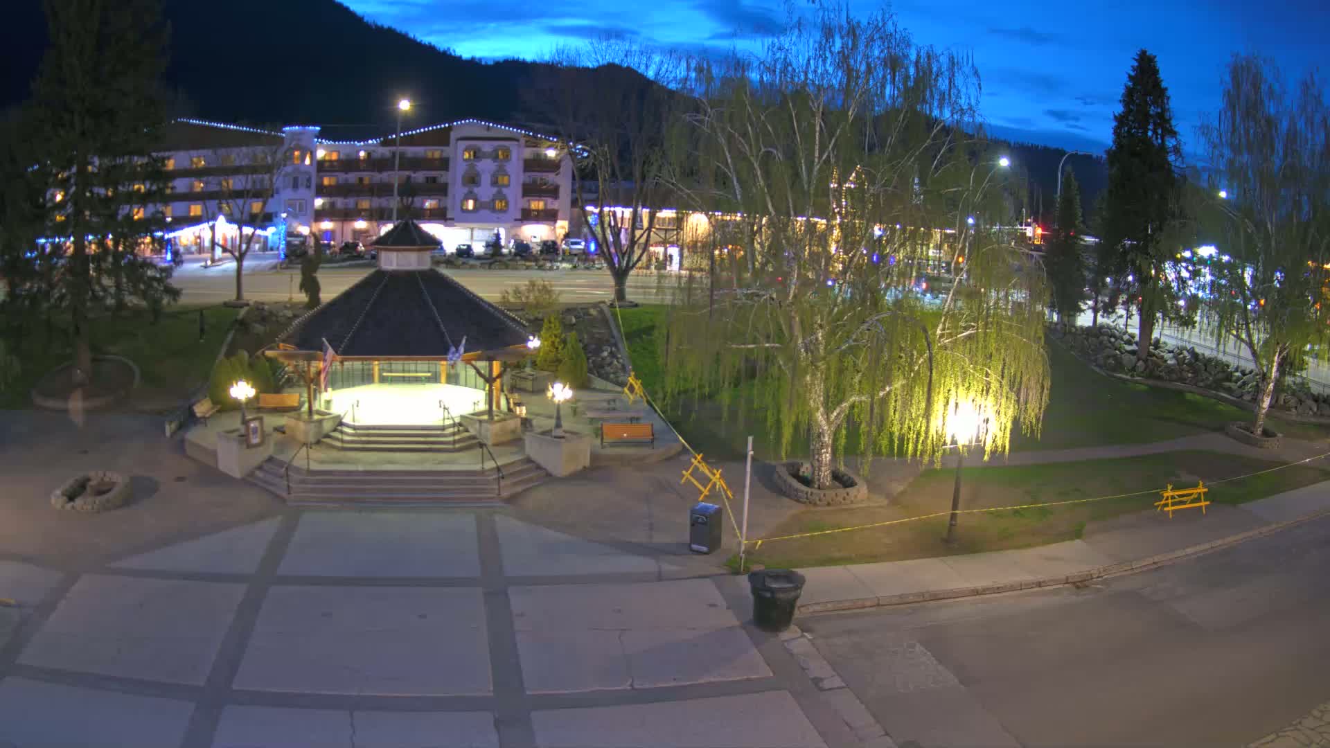 A nighttime view of a town square featuring a gazebo, trees, and a multi-story building in the background under a twilight sky.