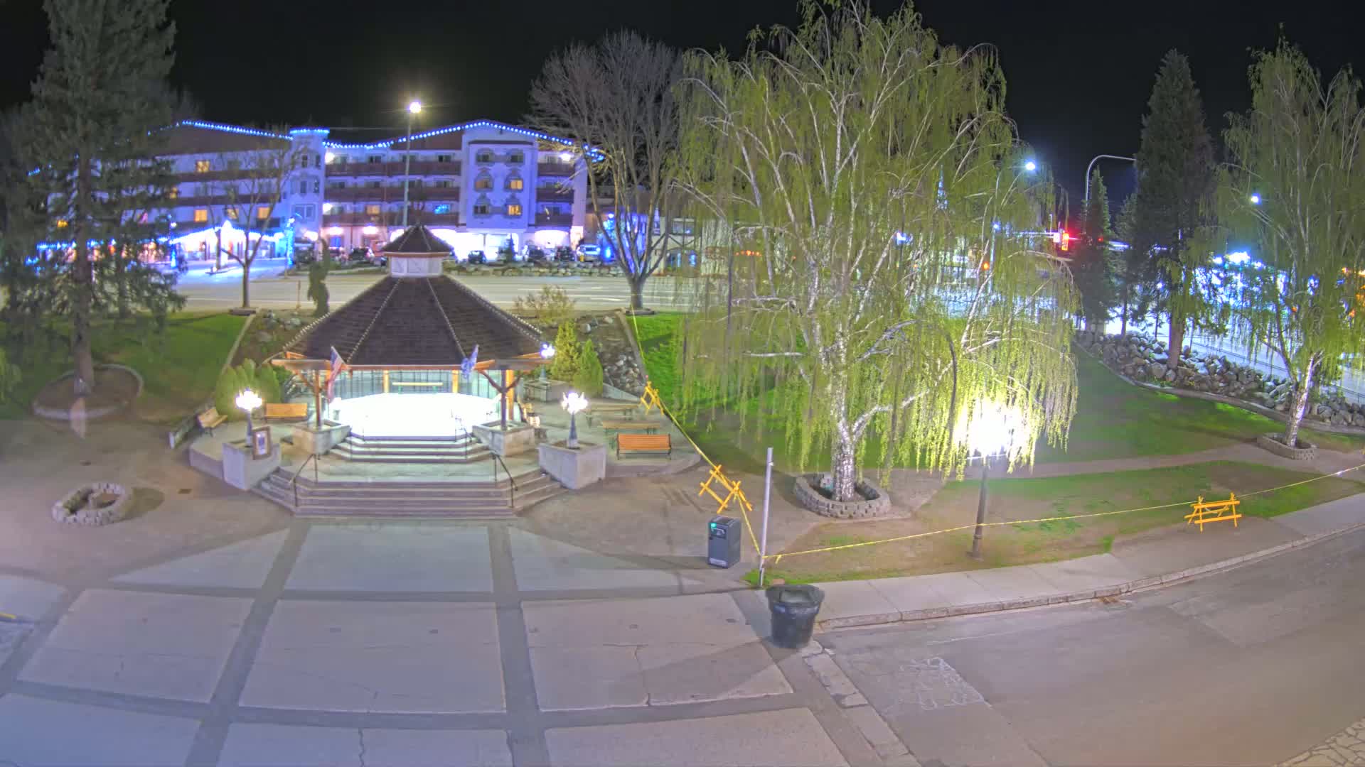A nighttime view of a town square featuring a gazebo, park benches, trees, and a multi-story building in the background, illuminated by streetlights.