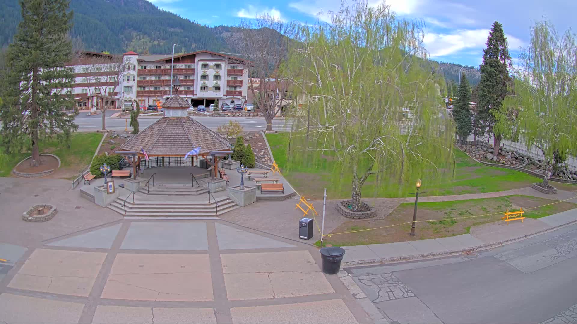 A wood-shingled gazebo sits in a paved plaza next to a grassy park, with a multi-story building and mountains in the background under a partly cloudy sky.