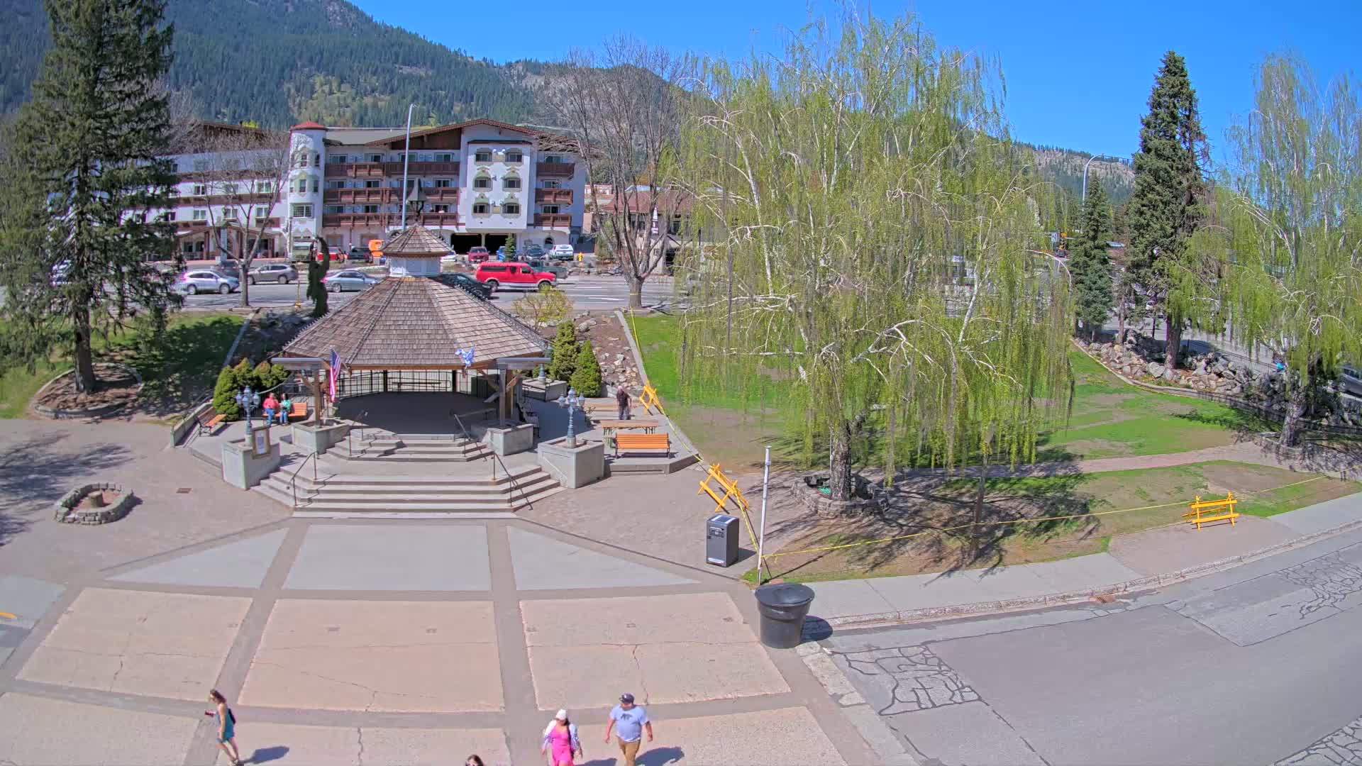 A paved plaza with a gazebo, benches, and several people walking, situated in front of a multi-story building, all under a sunny sky with mountains in the background.