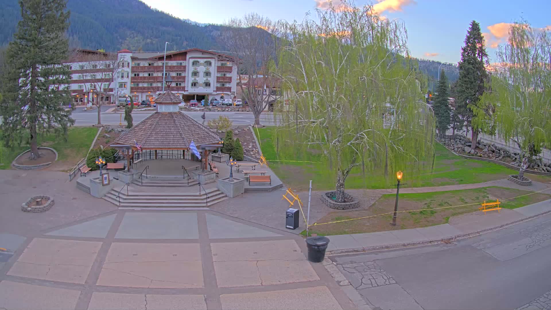 A gazebo sits in a town square next to a multi-story hotel, under a partly cloudy evening sky, with mountains in the background.