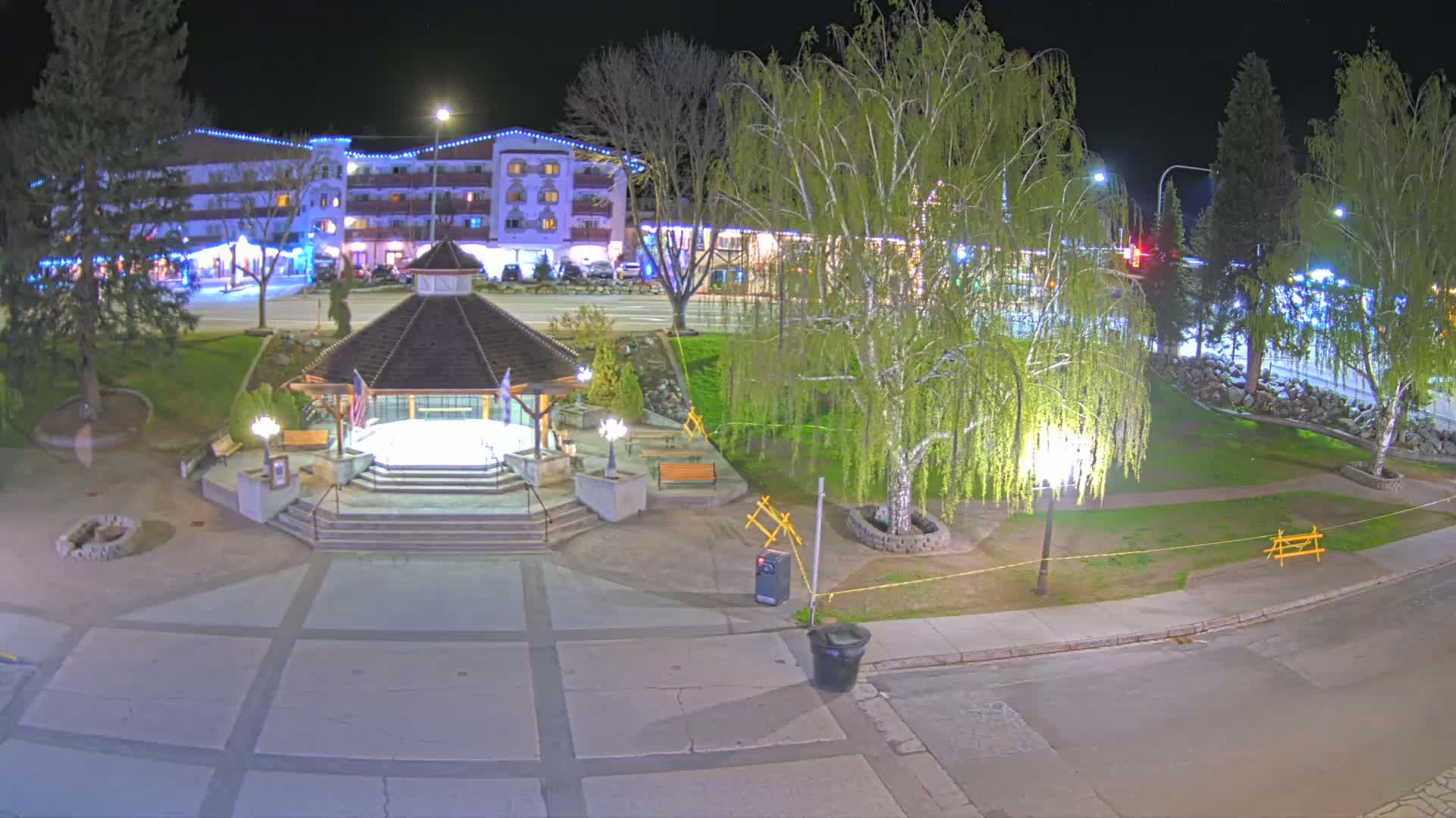 A nighttime view of a town square with a gazebo, trees, and a hotel in the background under clear skies.