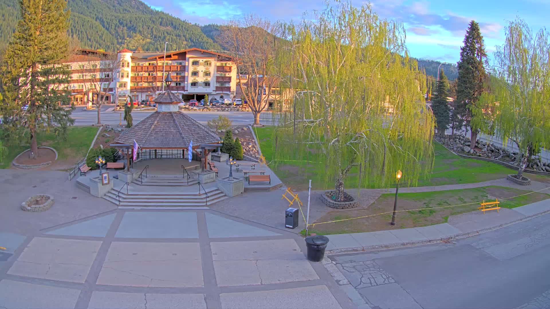 A gazebo sits in a paved plaza in front of a large hotel, with a grassy park and trees surrounding it under a partly cloudy sky.