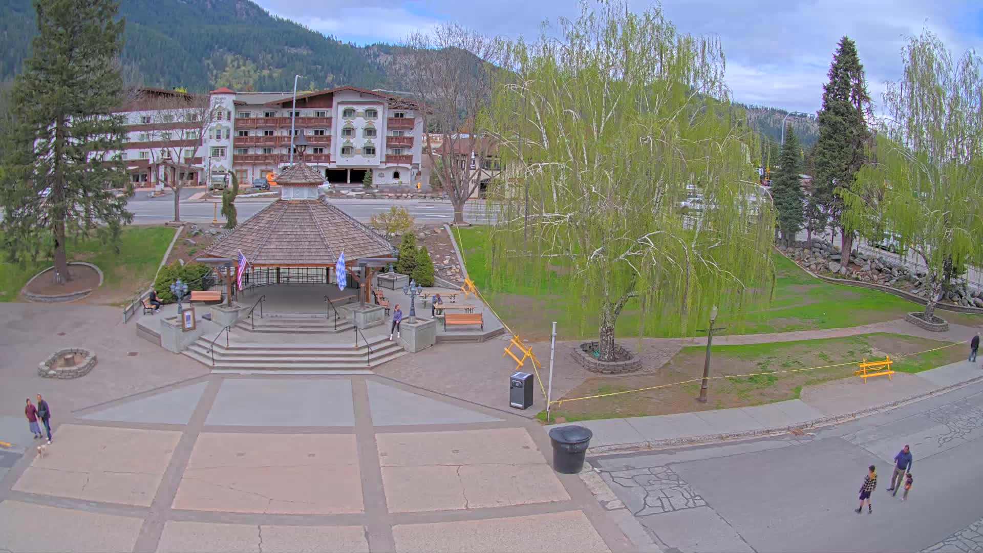 A paved plaza with a gazebo and several people, situated in front of a multi-story building, under a partly cloudy sky, is surrounded by green grass and trees; mountains are visible in the background.