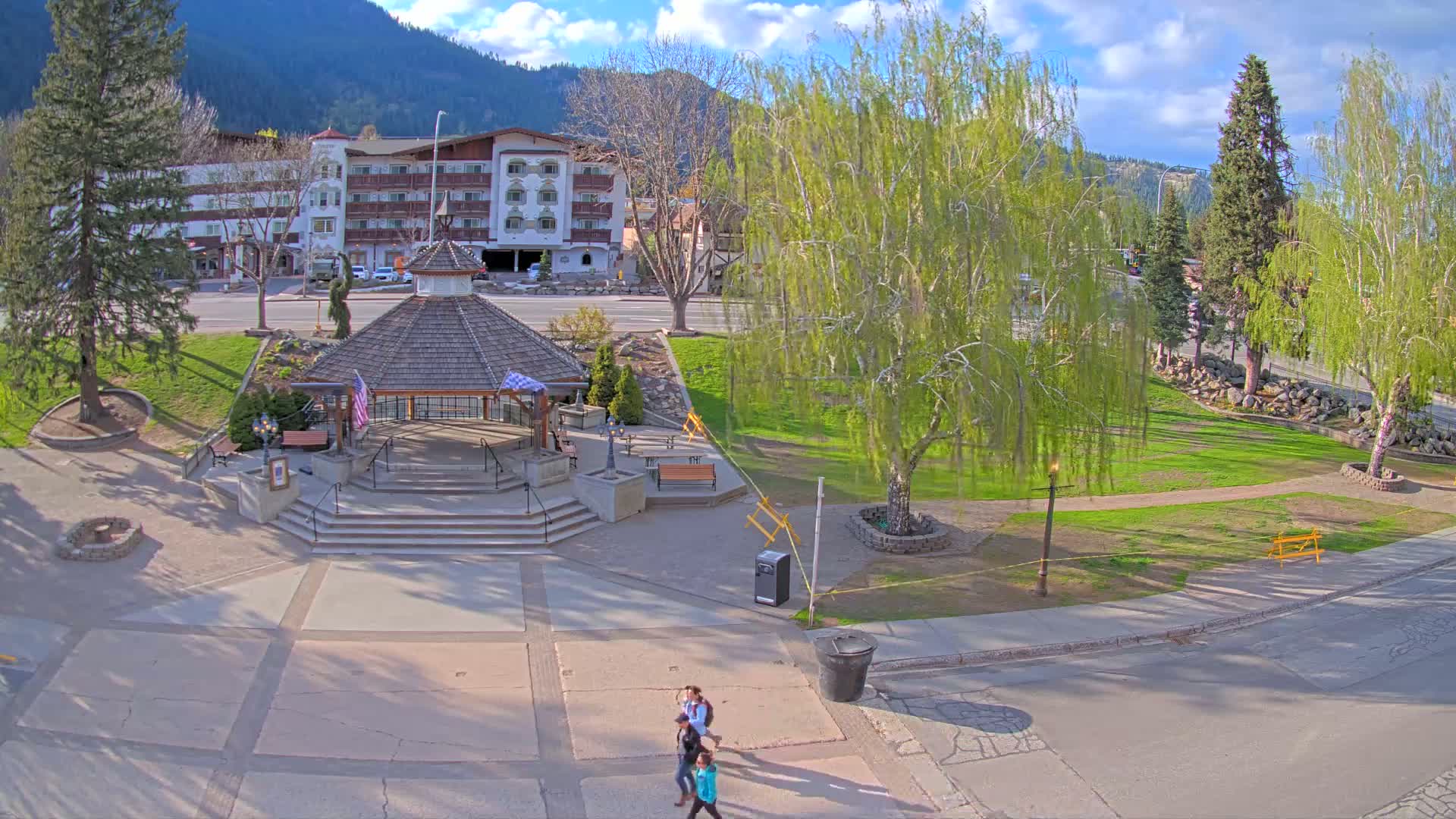 A park with a gazebo, green grass, and trees is situated in front of a multi-story building under a partly sunny sky.