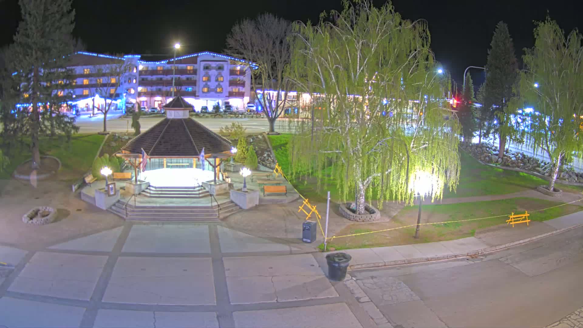 A nighttime view of a town square featuring a gazebo, landscaped areas, a multi-story building, and trees illuminated by streetlights.