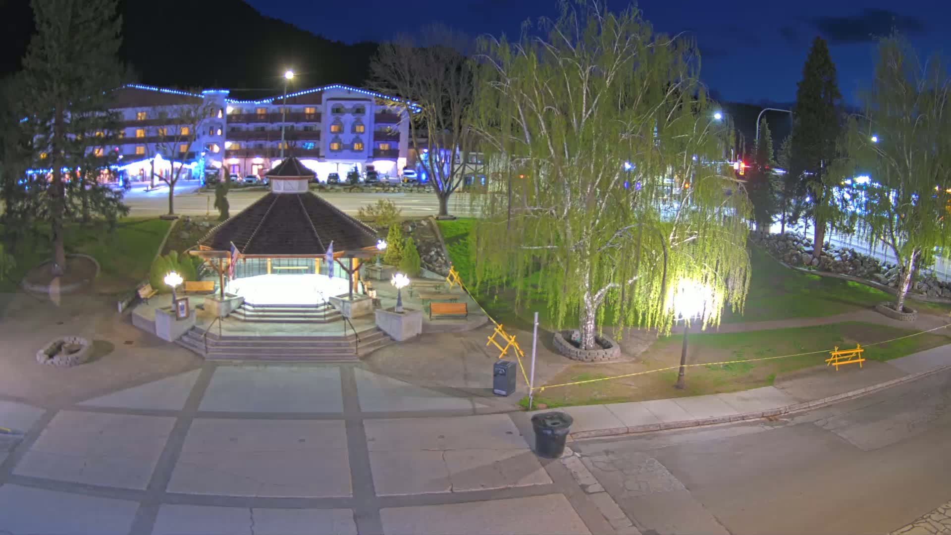 A nighttime view of a town square featuring a gazebo, trees, a building with lights, and a paved area.