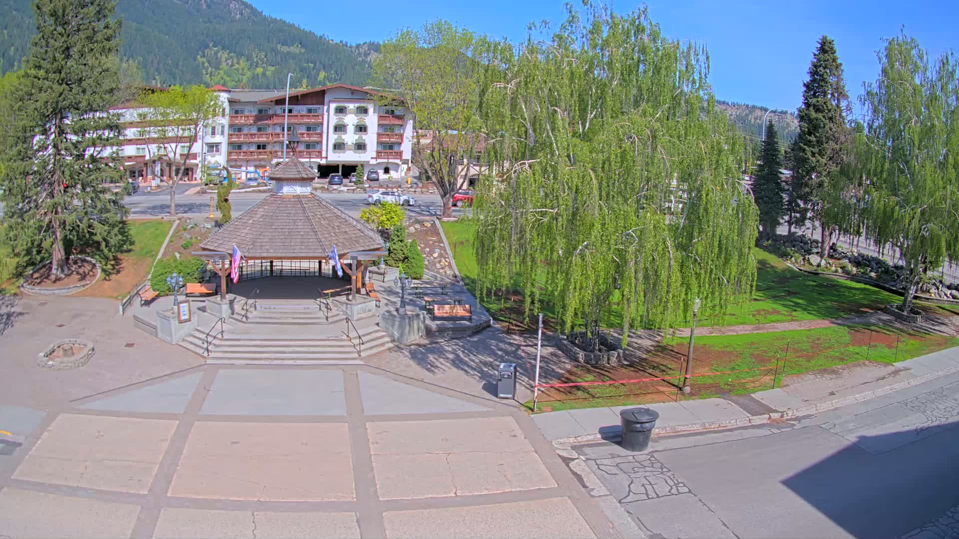 A paved plaza with a gazebo sits in front of a multi-story building, surrounded by green grass, trees, and mountains under a clear, sunny sky.