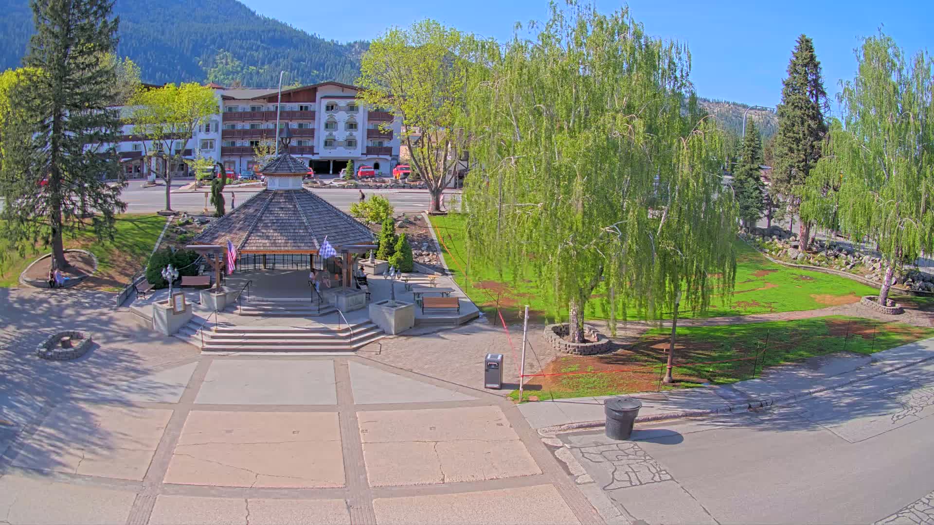 A gazebo sits in a paved plaza next to a grassy park with trees, under a sunny sky in front of a multi-story building and mountains in the background.