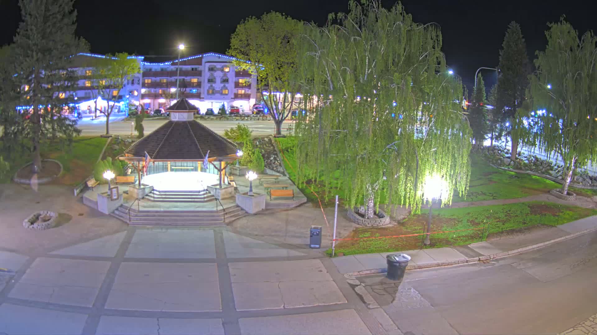 A nighttime view of a town square with a gazebo, trees, and a multi-story building in the background.