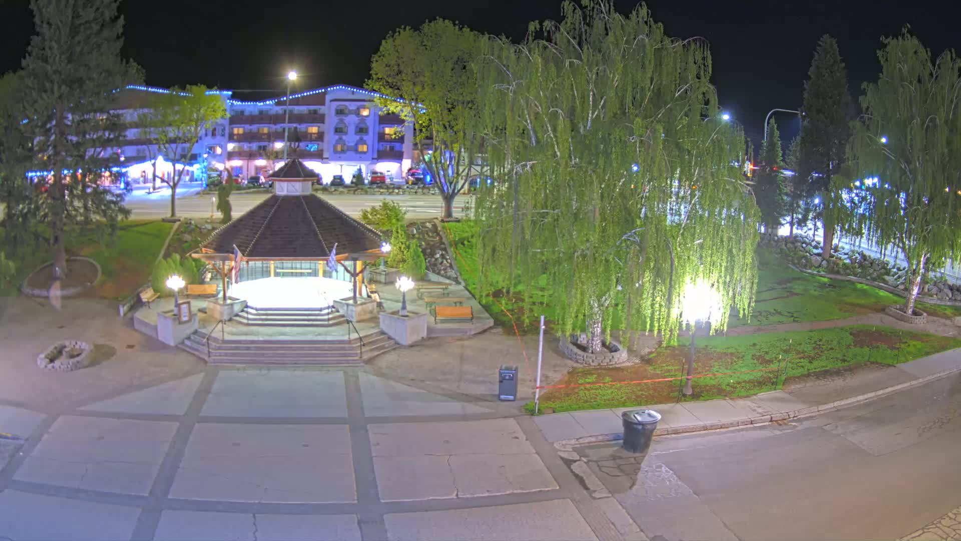 A nighttime view of a park with a gazebo, trees, and a building in the background, illuminated by streetlights.