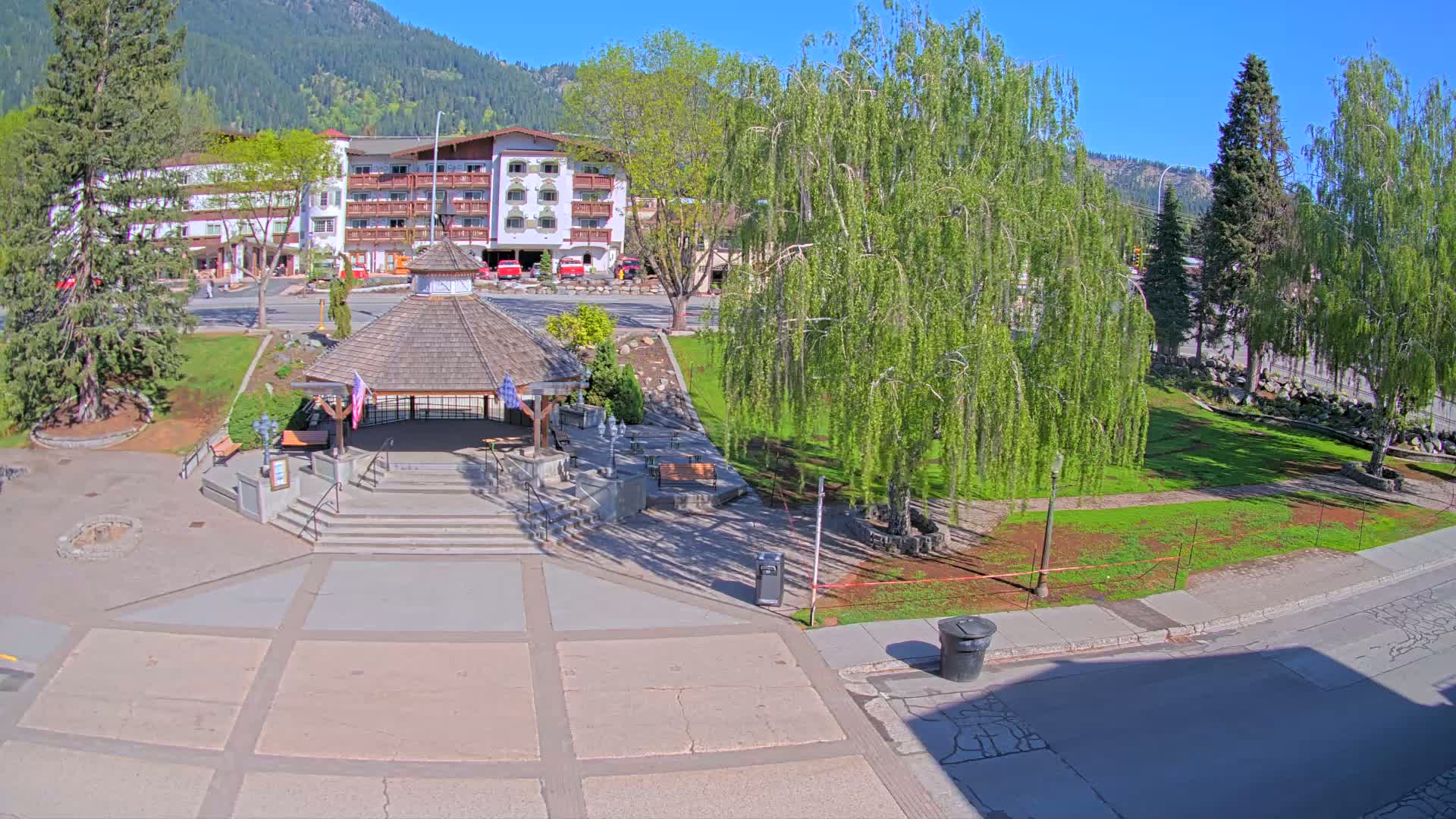 A paved plaza with a gazebo, grassy areas, trees, and a multi-story building in the background on a sunny day.