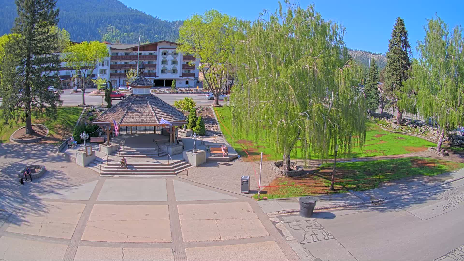 A town square with a gazebo, benches, and trees, situated in front of a multi-story building, on a sunny day with mountains in the background.