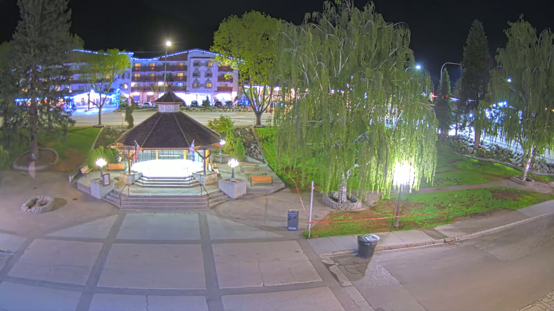 A nighttime view of a park with a gazebo, trees, and a hotel in the background, illuminated by streetlights.