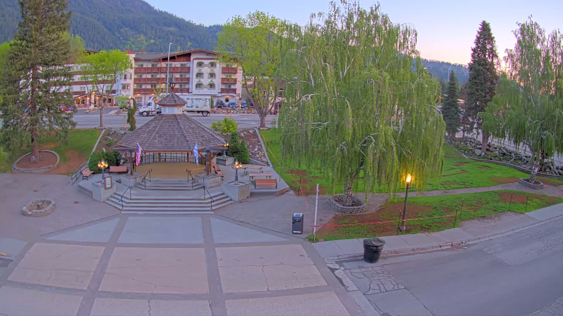 A paved plaza with a gazebo, surrounded by grass, trees, and a multi-story building, in the early evening.
