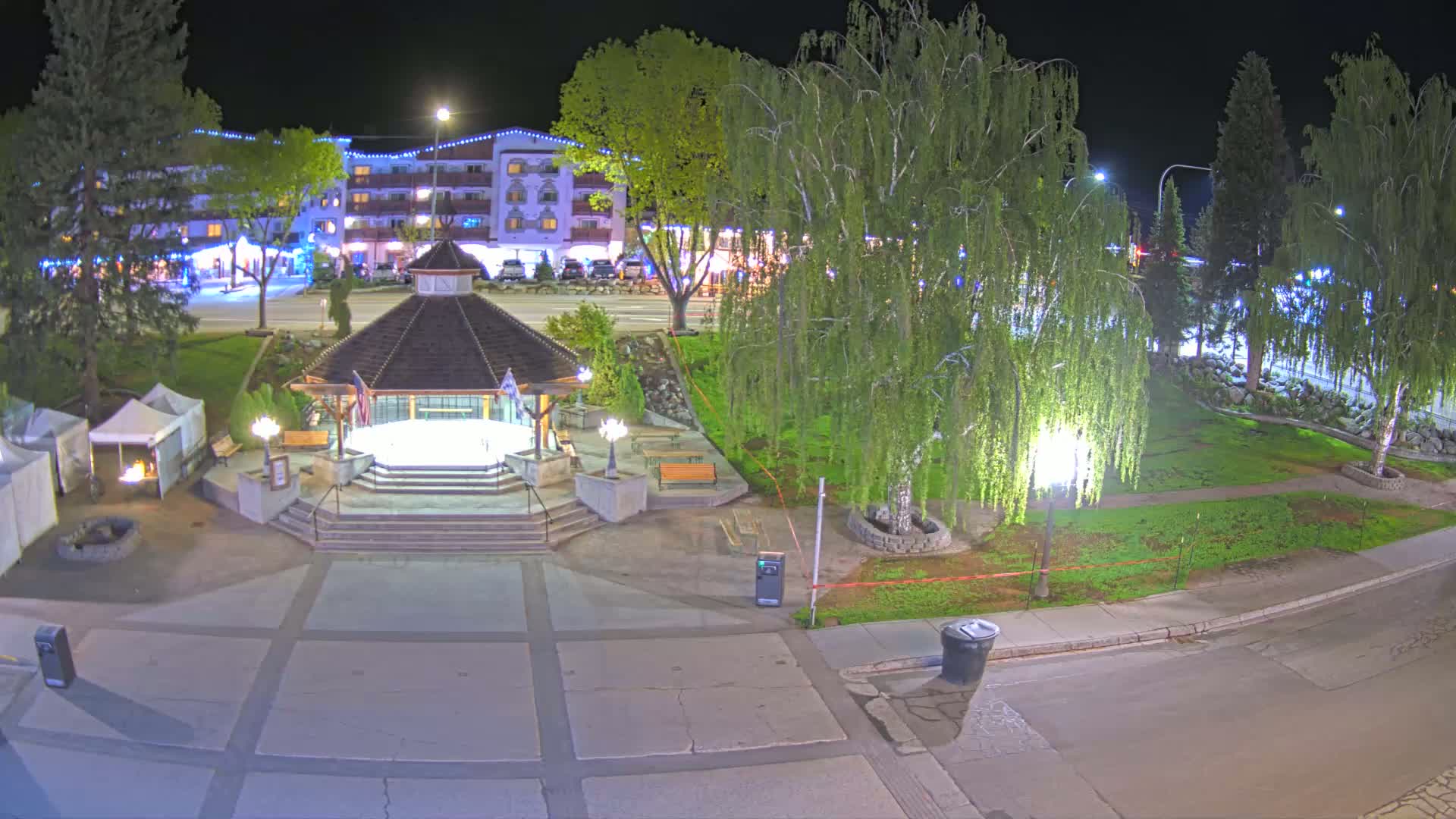 A nighttime view of a town square featuring a gazebo, landscaping, and a building in the background under clear skies.