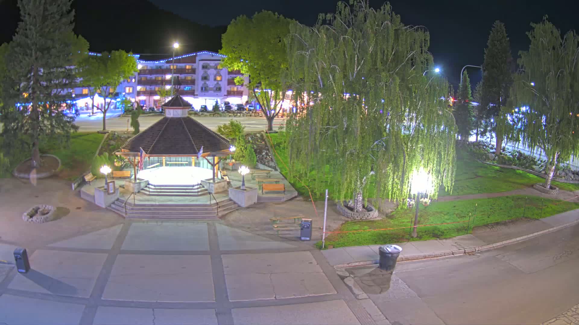 A nighttime view of a town square featuring a gazebo, landscaping, and a multi-story building in the background.