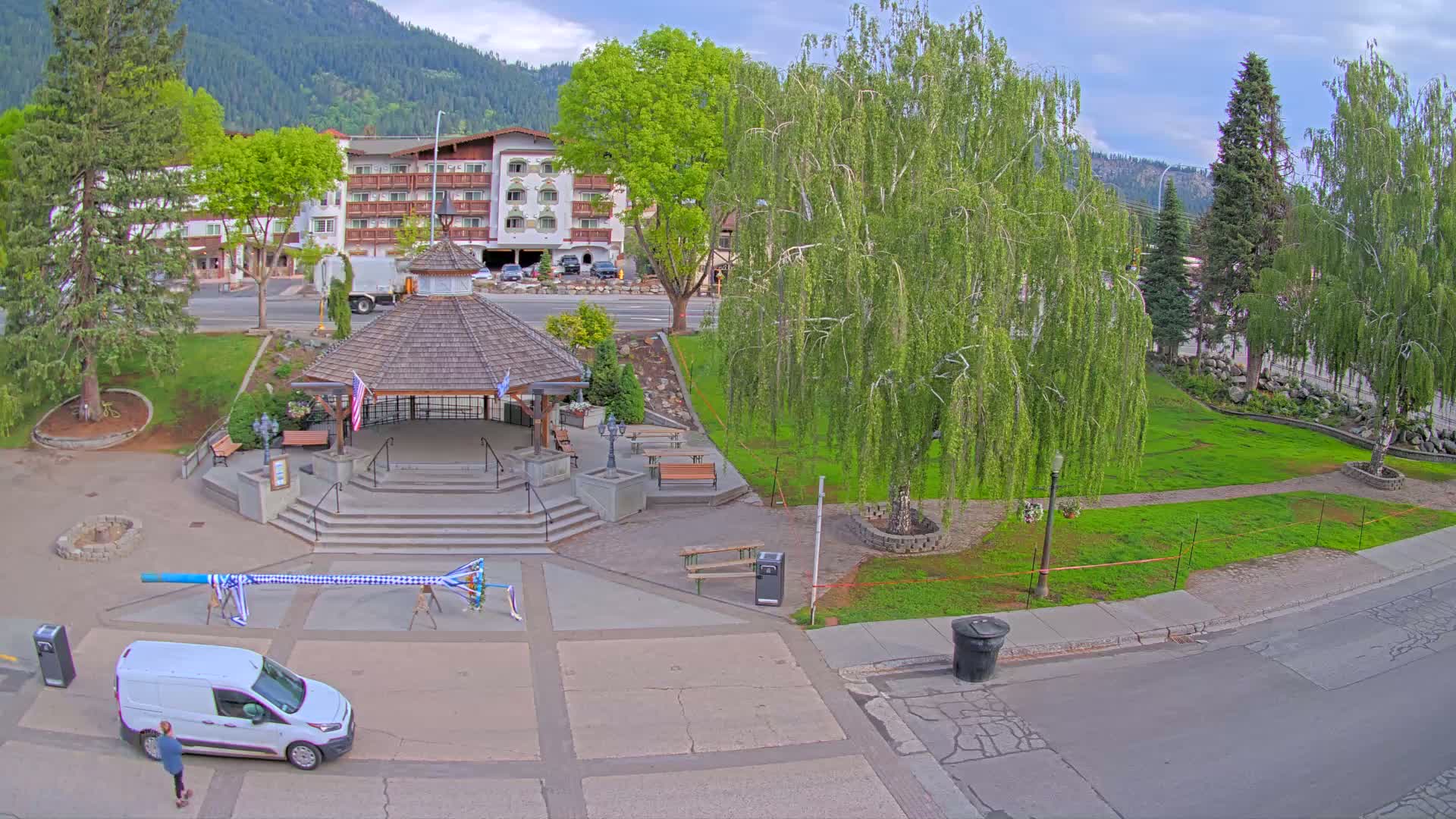 A park with a gazebo, a long maypole, and a multi-story building in the background under partly cloudy skies.
