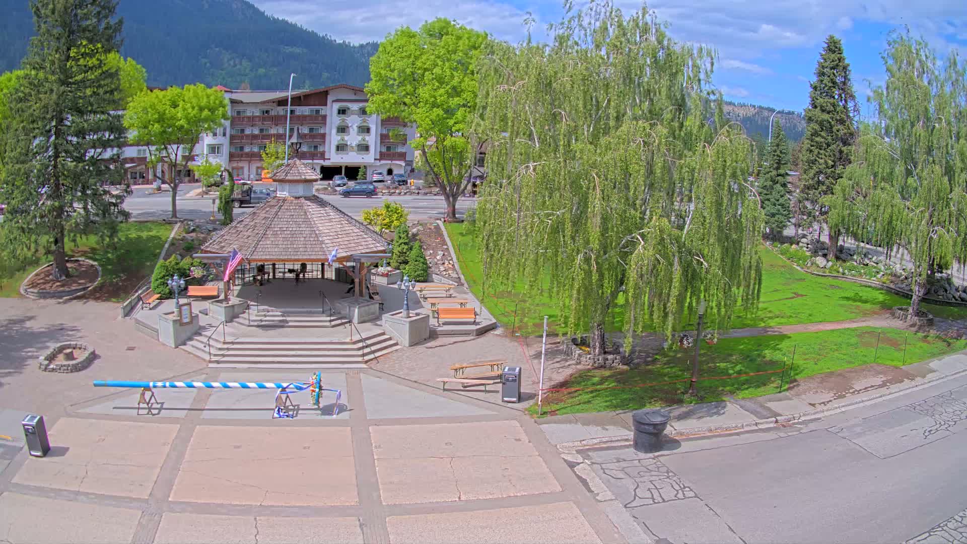 A park with a gazebo, picnic tables, and a large maypole, situated next to a multi-story building under a partly cloudy sky.