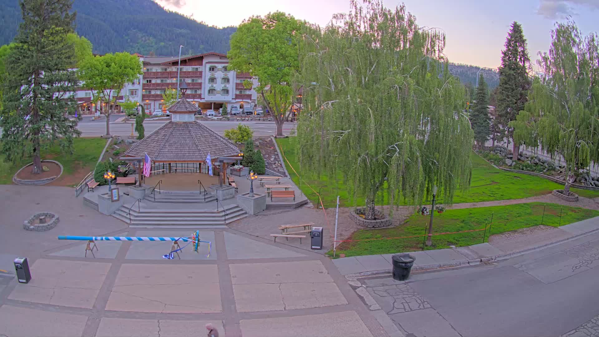 A large maypole stands in a town square next to a gazebo, with a hotel and mountains visible in the background under a twilight sky.