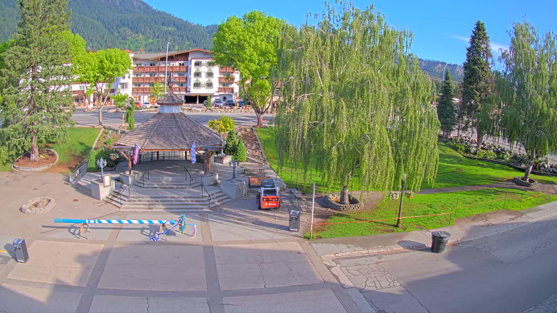 A town square with a gazebo, landscaping, a multi-story building in the background, and a maypole, all under a sunny sky.