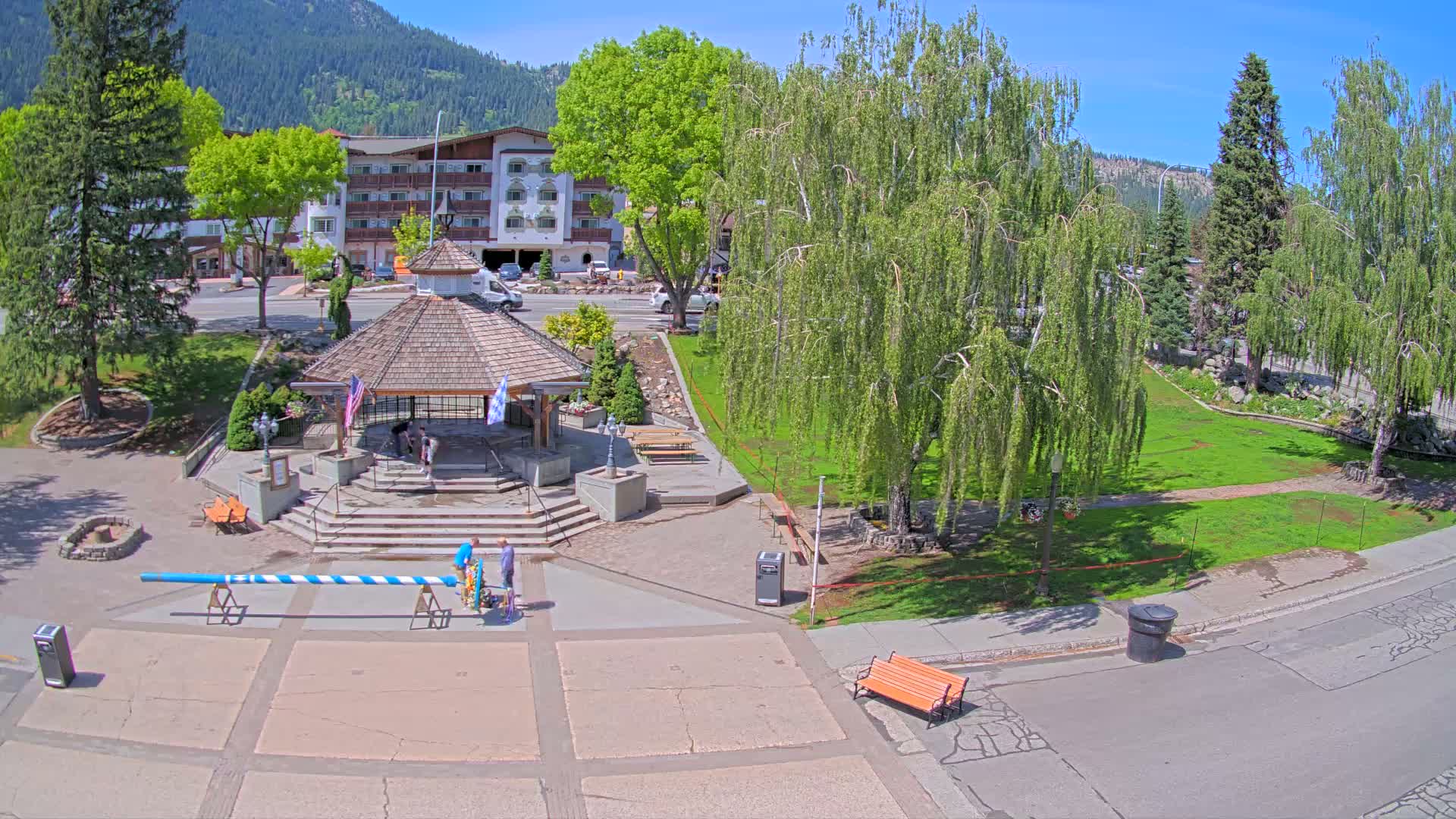 A paved town square with a gazebo, several trees, a long Maypole, and a few people, all under a sunny sky and mountains in the background.