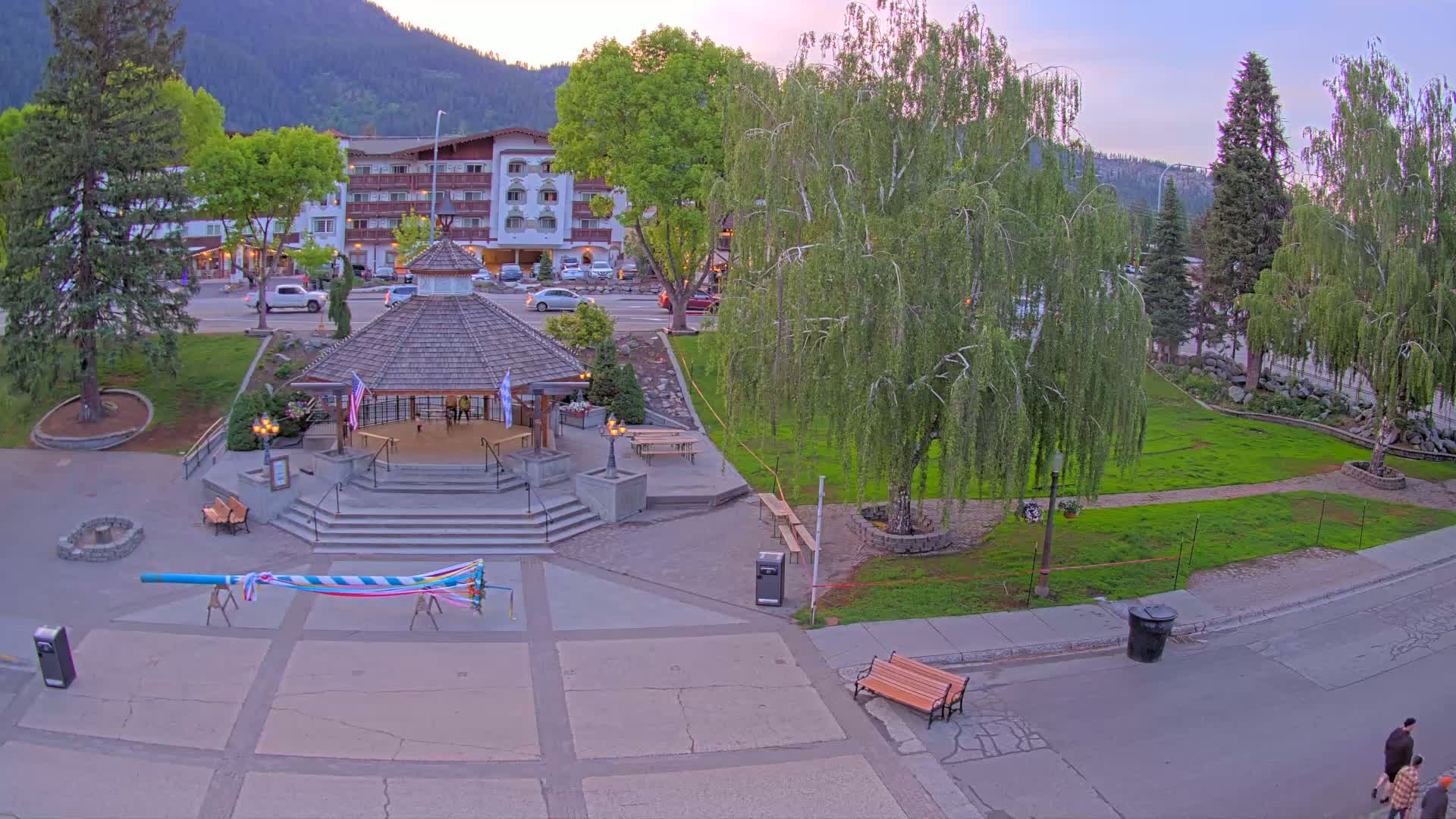A gazebo sits in a town square with a grassy area, trees, and a multi-story building in the background under a partly cloudy evening sky.