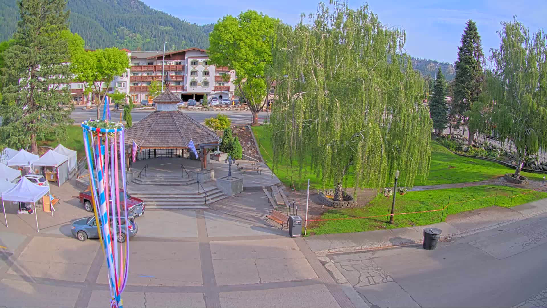 A town square with a gazebo, several cars, a maypole, and lush green trees under a sunny sky, all situated in front of a multi-story building and mountains in the background.