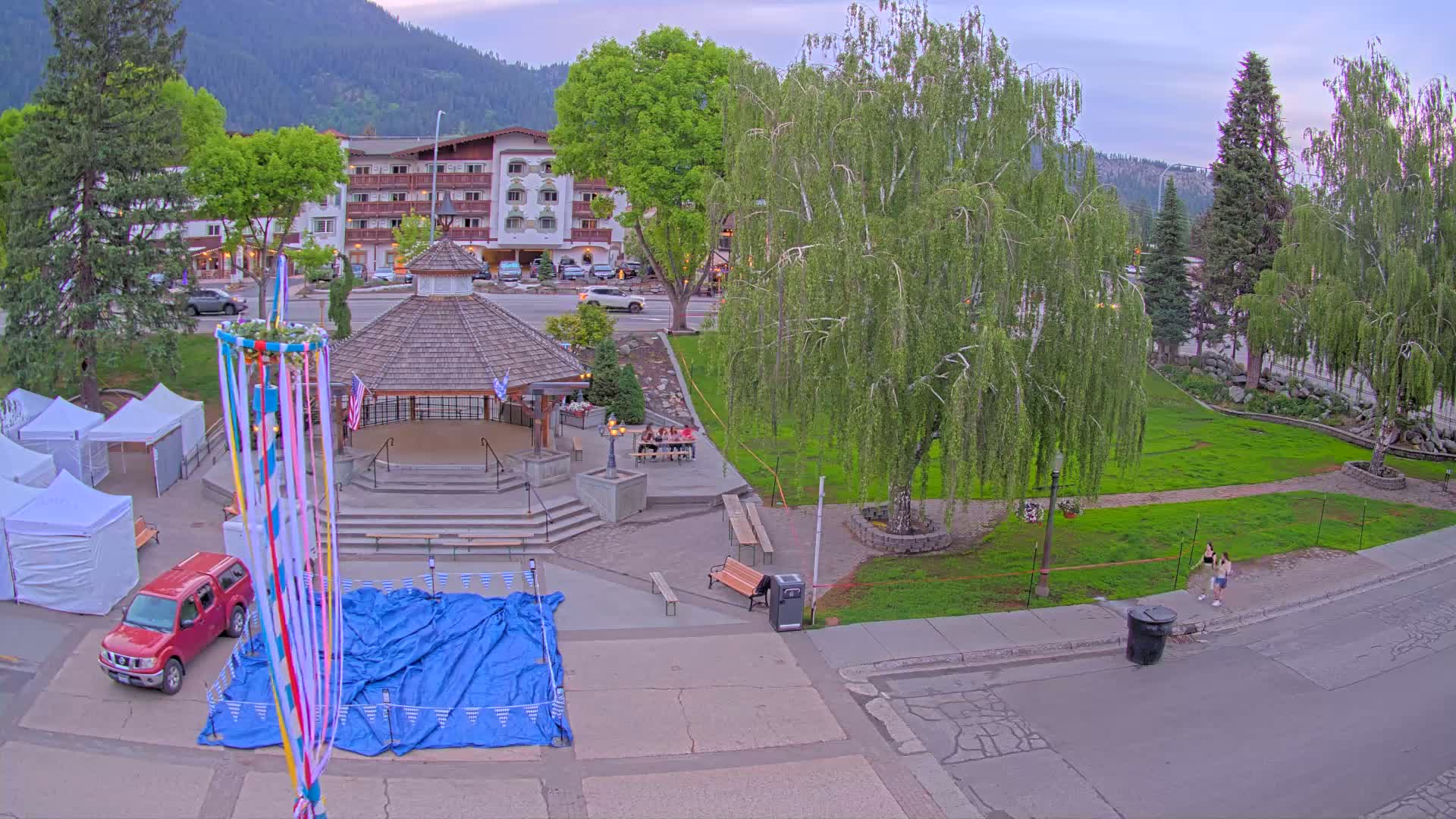 A town square with a gazebo, maypole, and several people, under a partly cloudy evening sky, features a grassy area with trees, some tents, and a red pickup truck parked nearby.