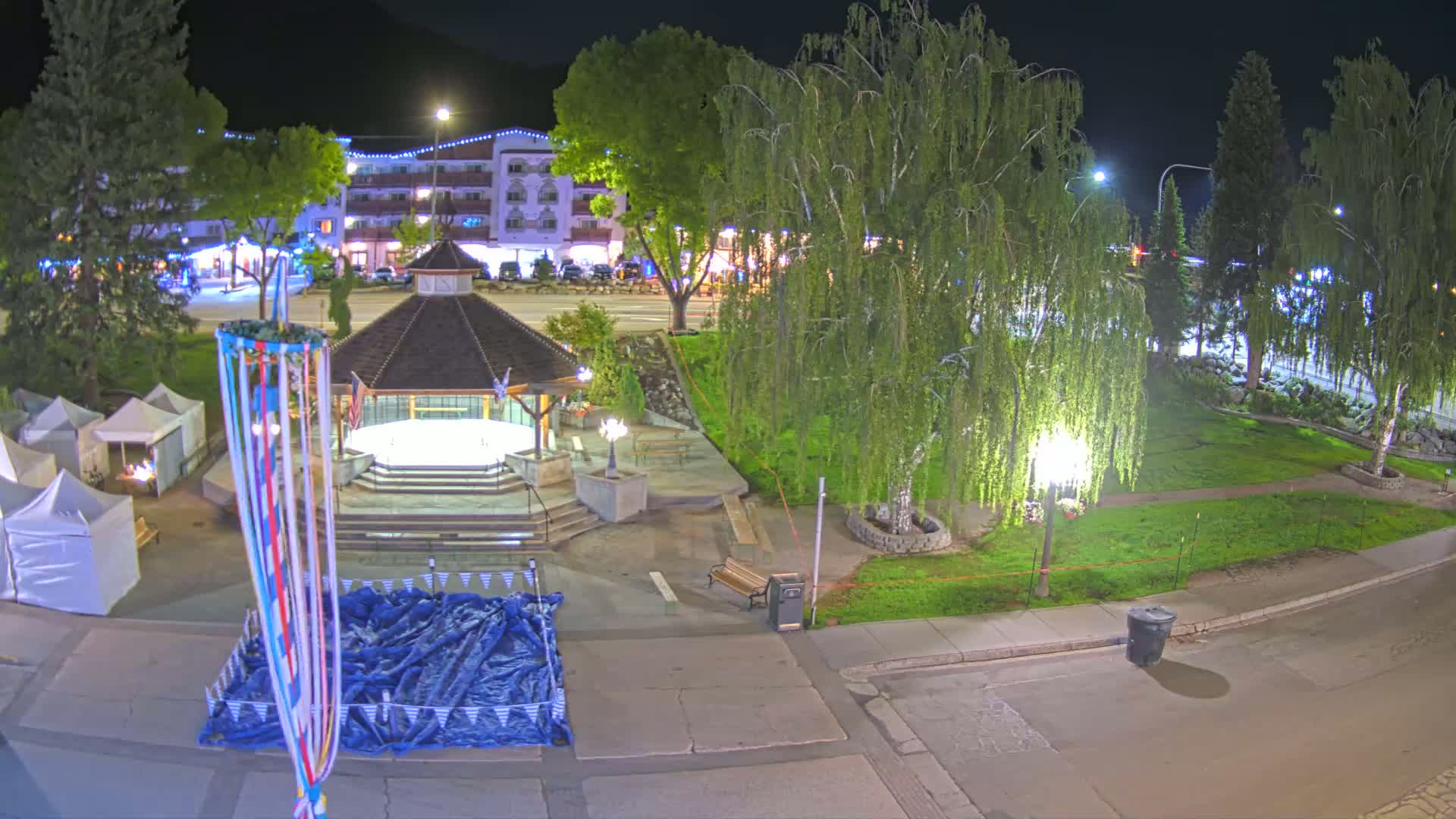 At night, a town square with a gazebo, a maypole, several tents, and trees is illuminated under artificial light.