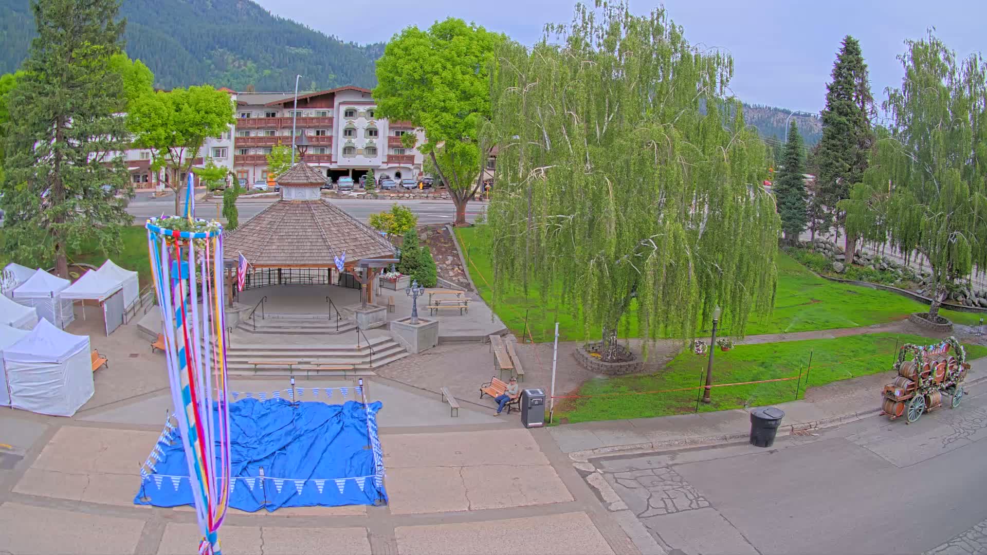 An overhead view of a town square with a gazebo, maypole, and several tents, under an overcast sky, next to a grassy area with trees and a multi-story building in the background.