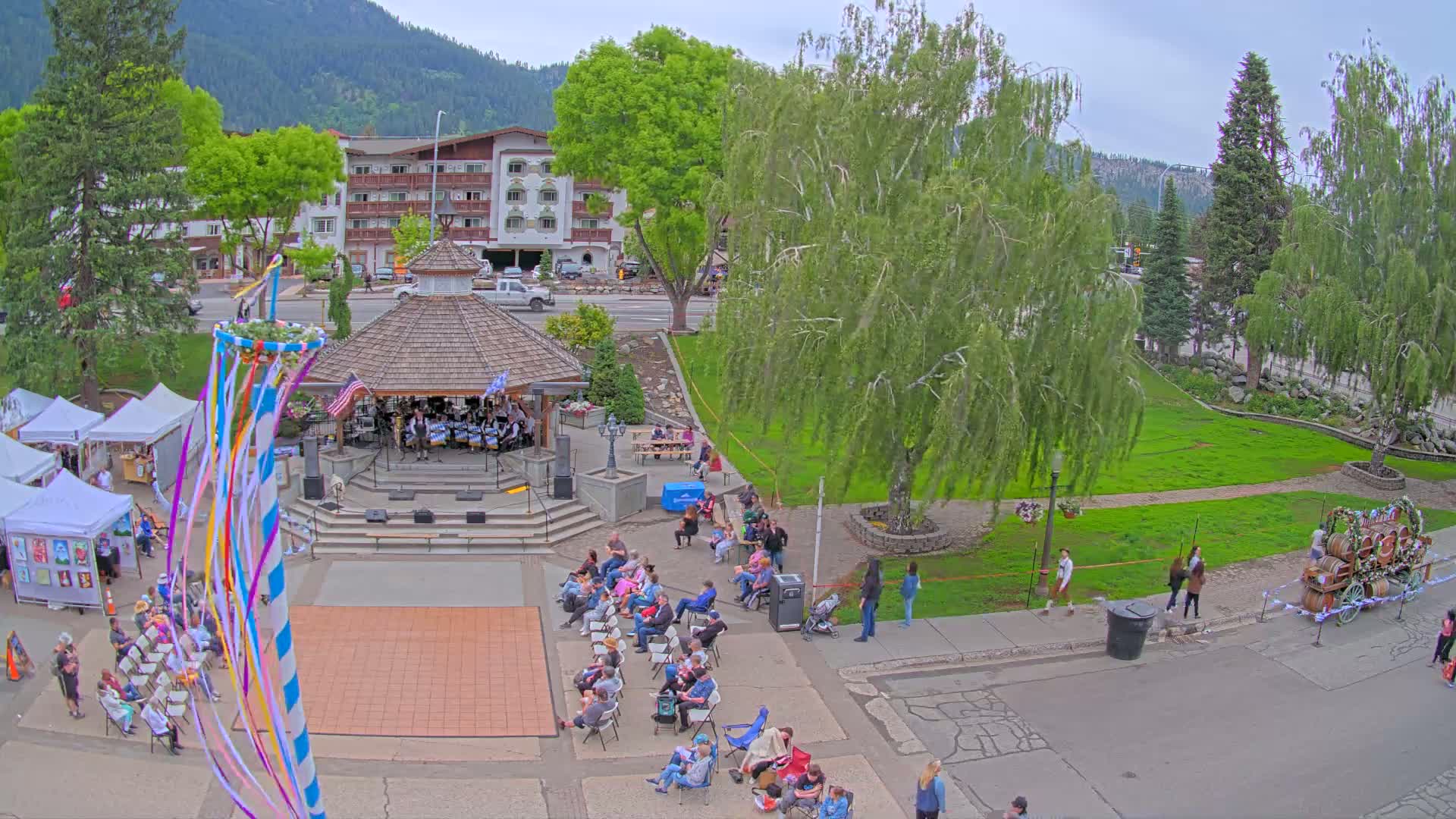 An outdoor festival scene under a cloudy sky features a band playing in a gazebo, people sitting in chairs, a maypole, and vendor tents.